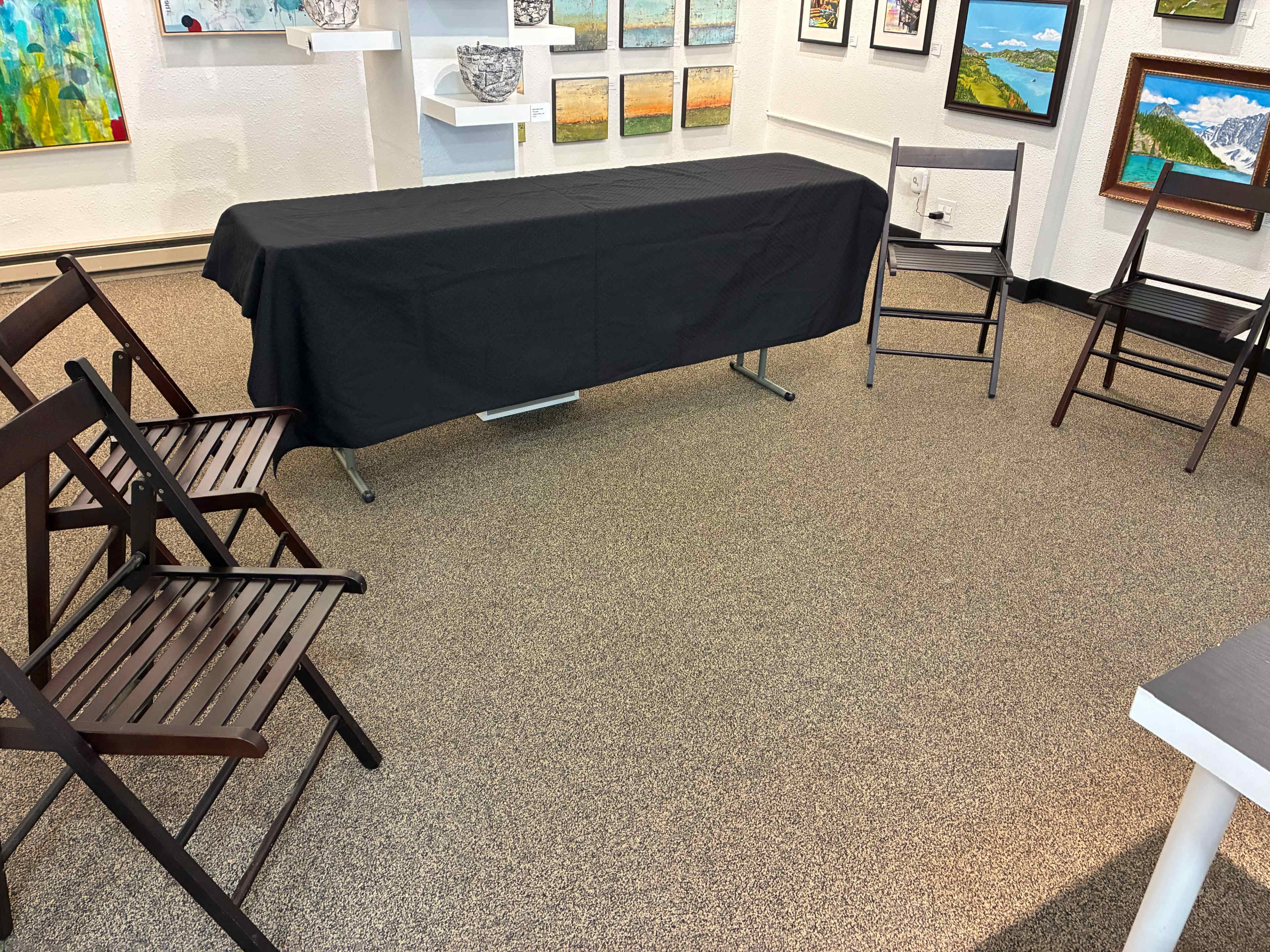 The image shows a room set up for an event, featuring a covered table in the center surrounded by several wooden chairs on a carpeted floor.