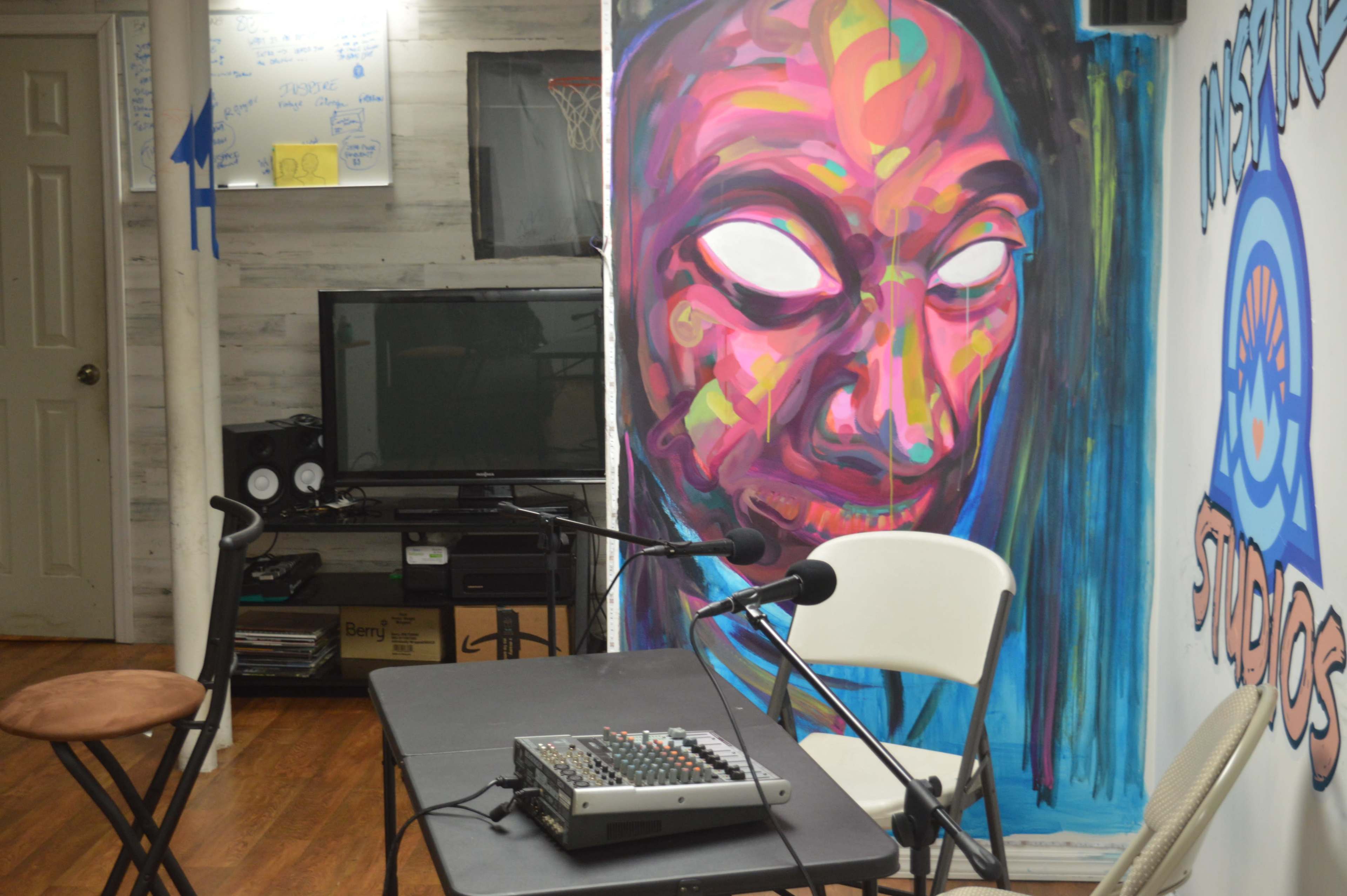 A small studio space features a table with a sound mixer, two microphones, and a colorful mural of a face on the wall beside a television.