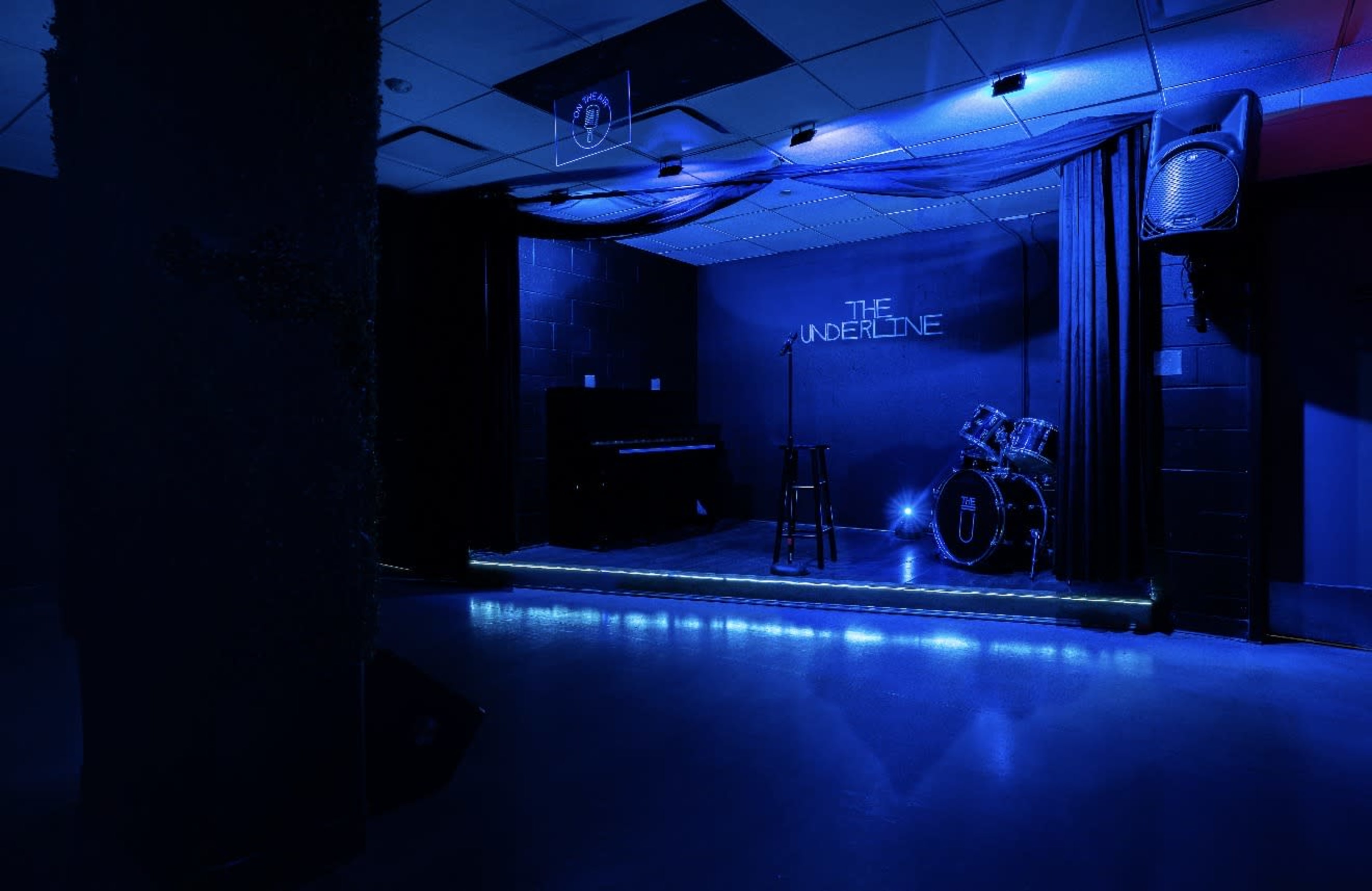 A dimly lit stage with blue lighting features a microphone stand, a drum set, and a piano in a performance space named "THE UNDERLINE."