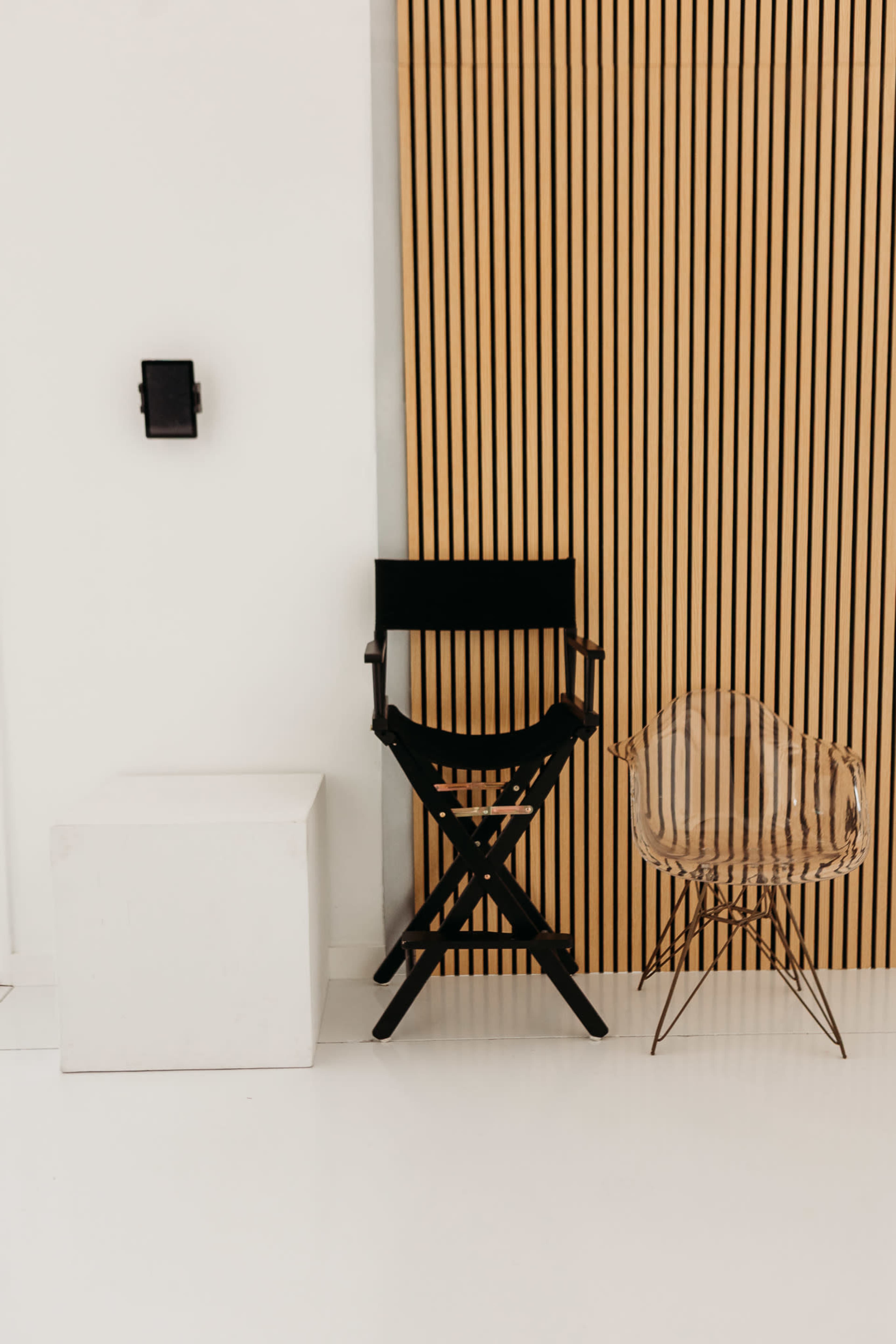 The image shows a black folding director's chair and a transparent acrylic chair next to a white cube against a backdrop of vertical wooden slats.