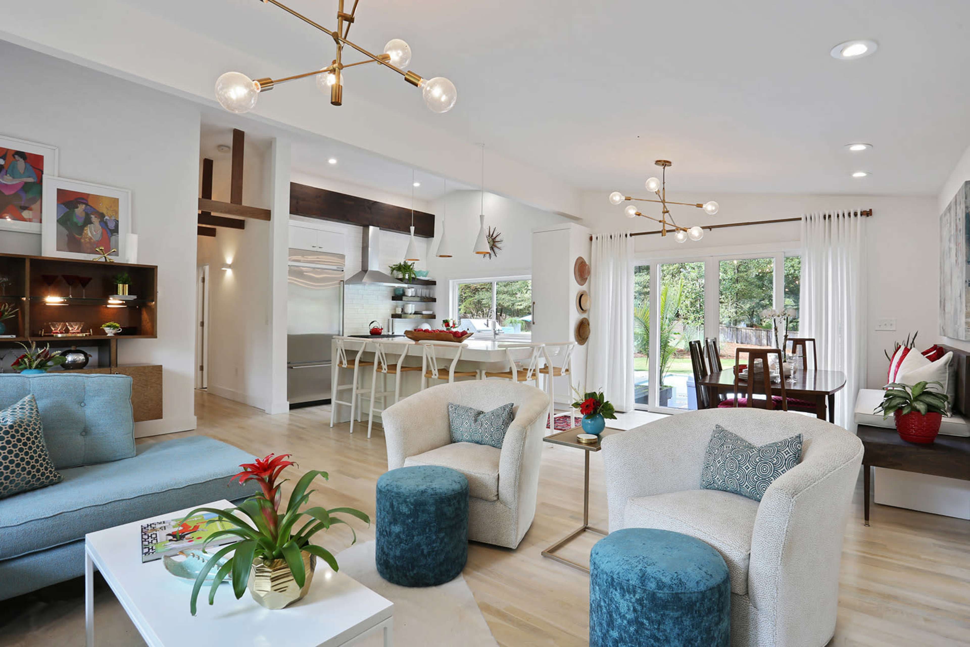 A modern living space features a seating area with light-colored furniture, a dining table in the background, and large windows that open to a view of greenery.