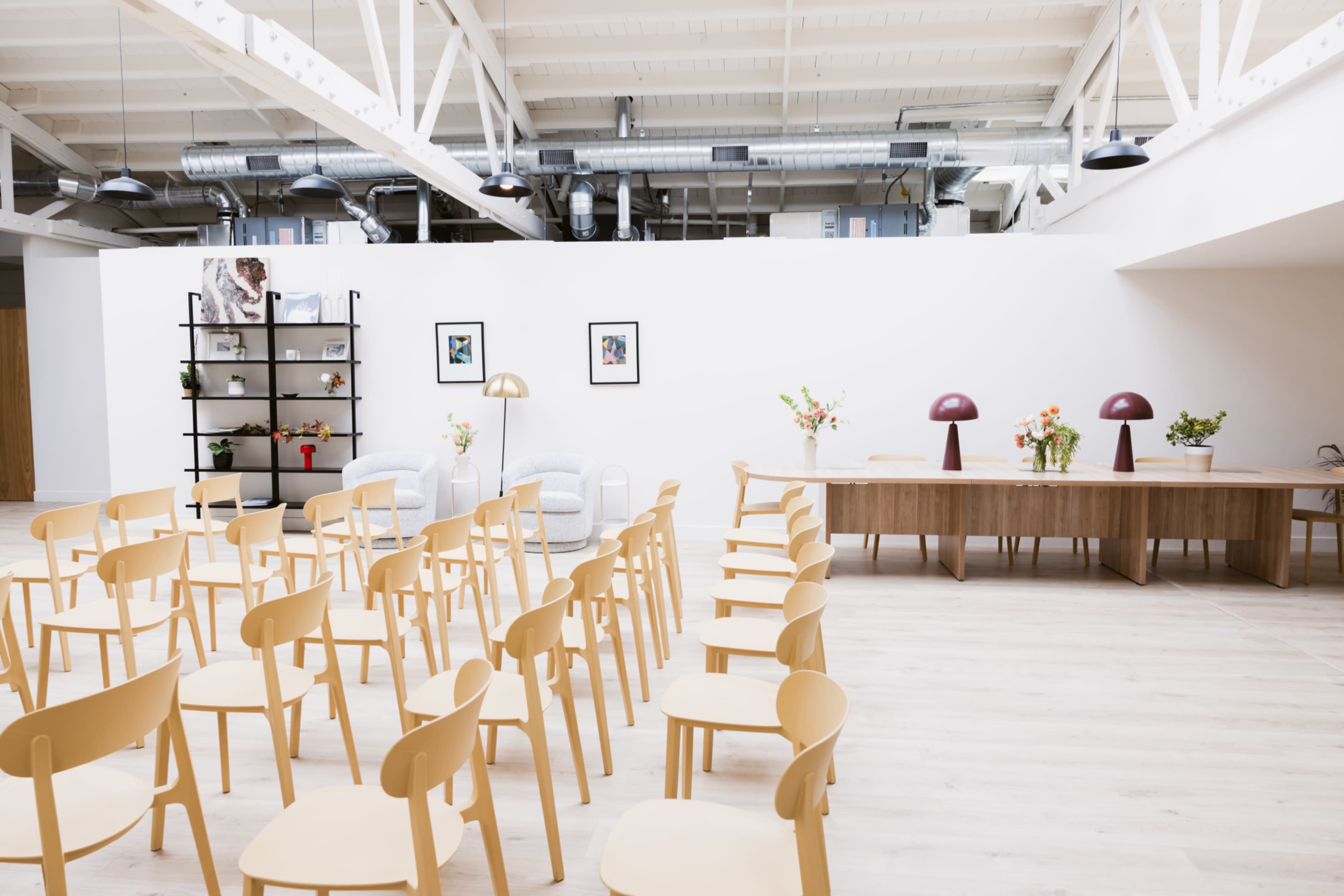 A spacious, minimalistic room features rows of empty wooden chairs facing a long table, with decorative elements like flowers and framed photos along the wall.