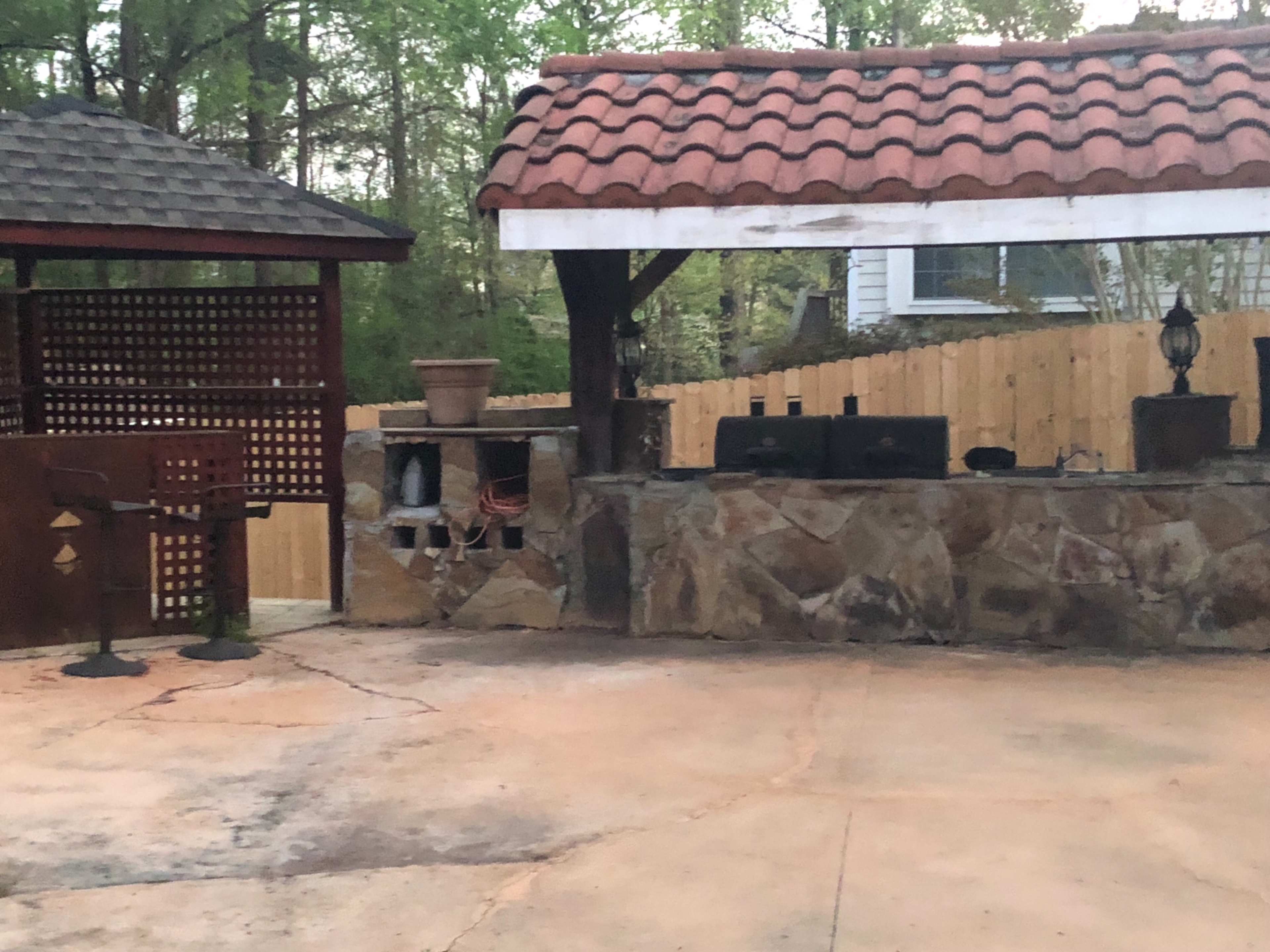 An outdoor patio area with a stone barbecue setup, a wooden gazebo, and a fenced background.