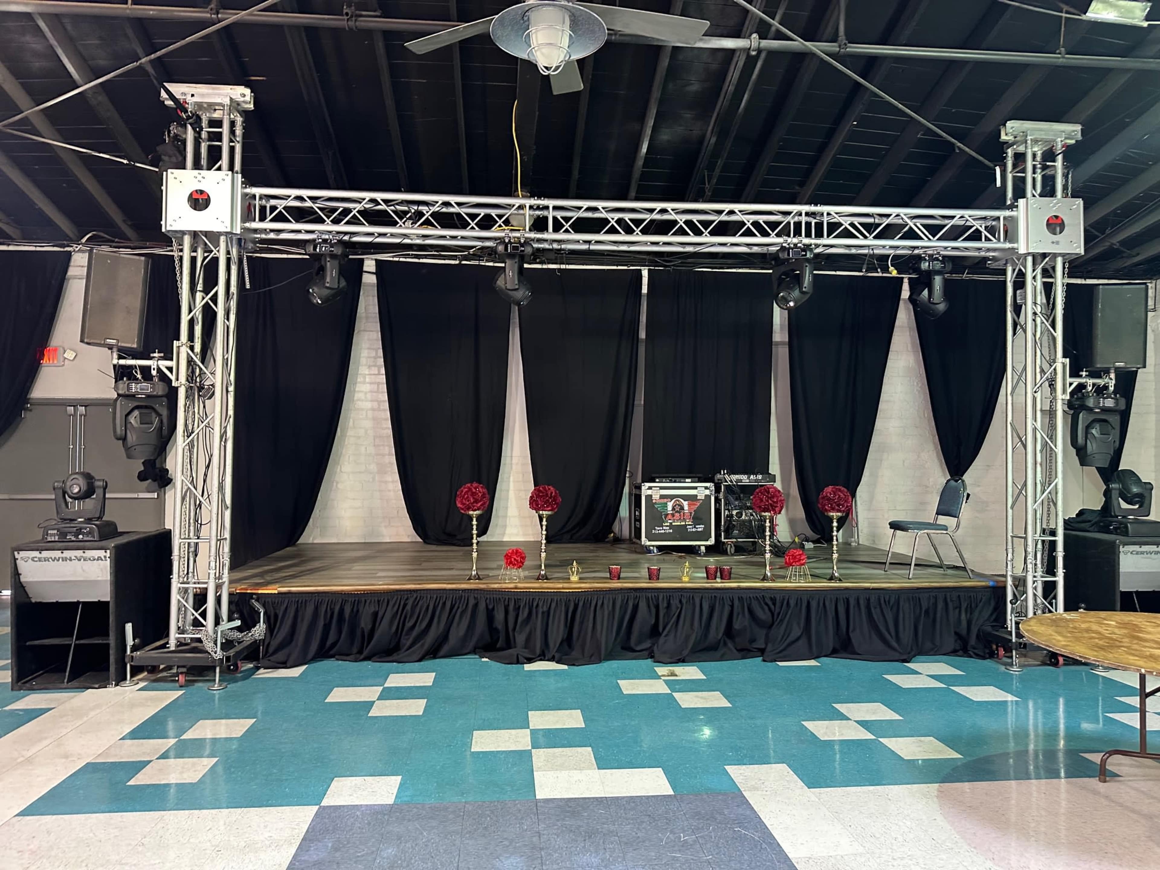 An empty stage with a backdrop of black drapes, flanked by tall speakers and adorned with red floral arrangements and candles on a wooden platform.