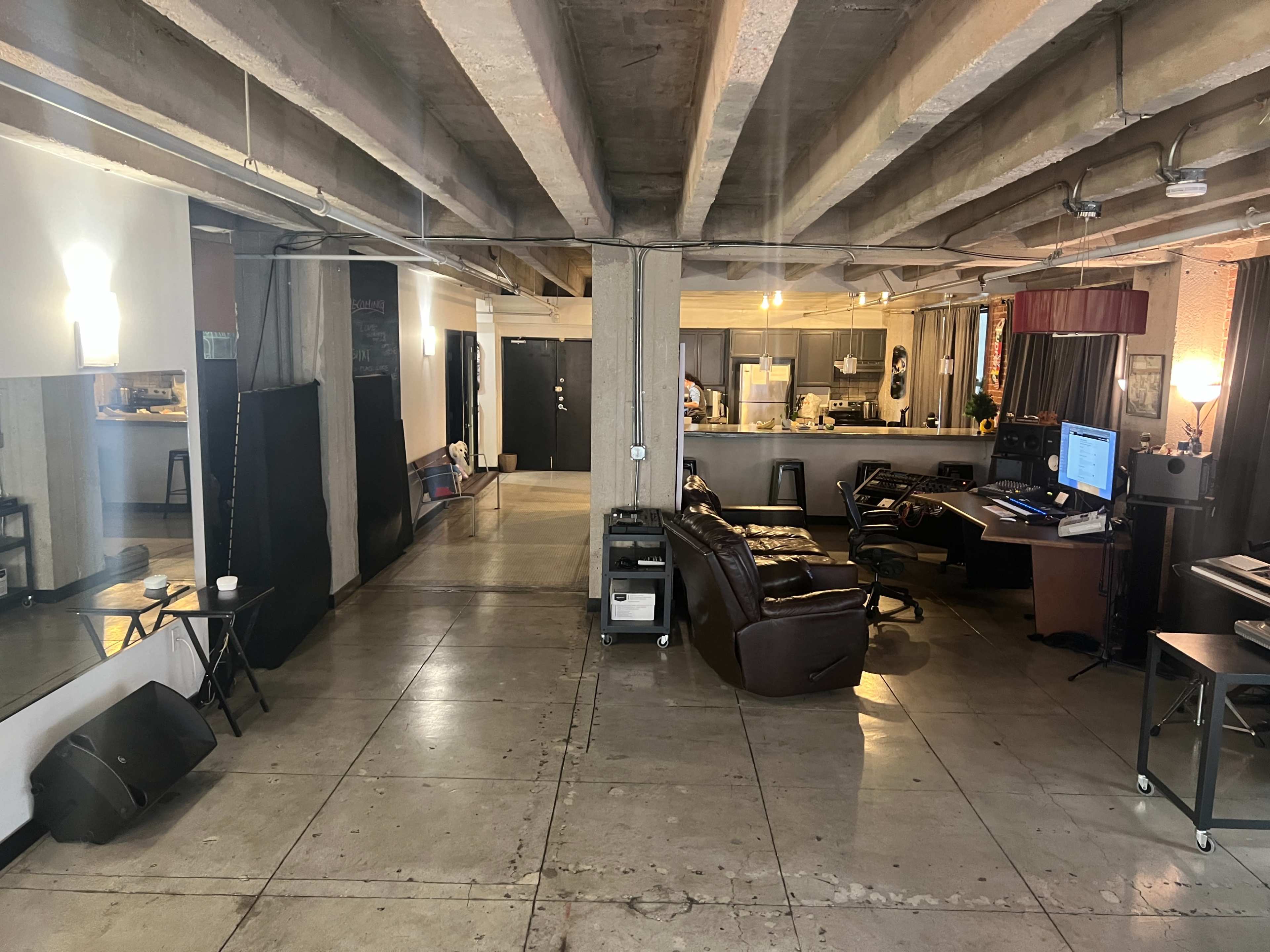 The image shows an open, spacious room with concrete floors, visible supports overhead, and areas designated for both living and working, including a kitchen and seating arrangement.