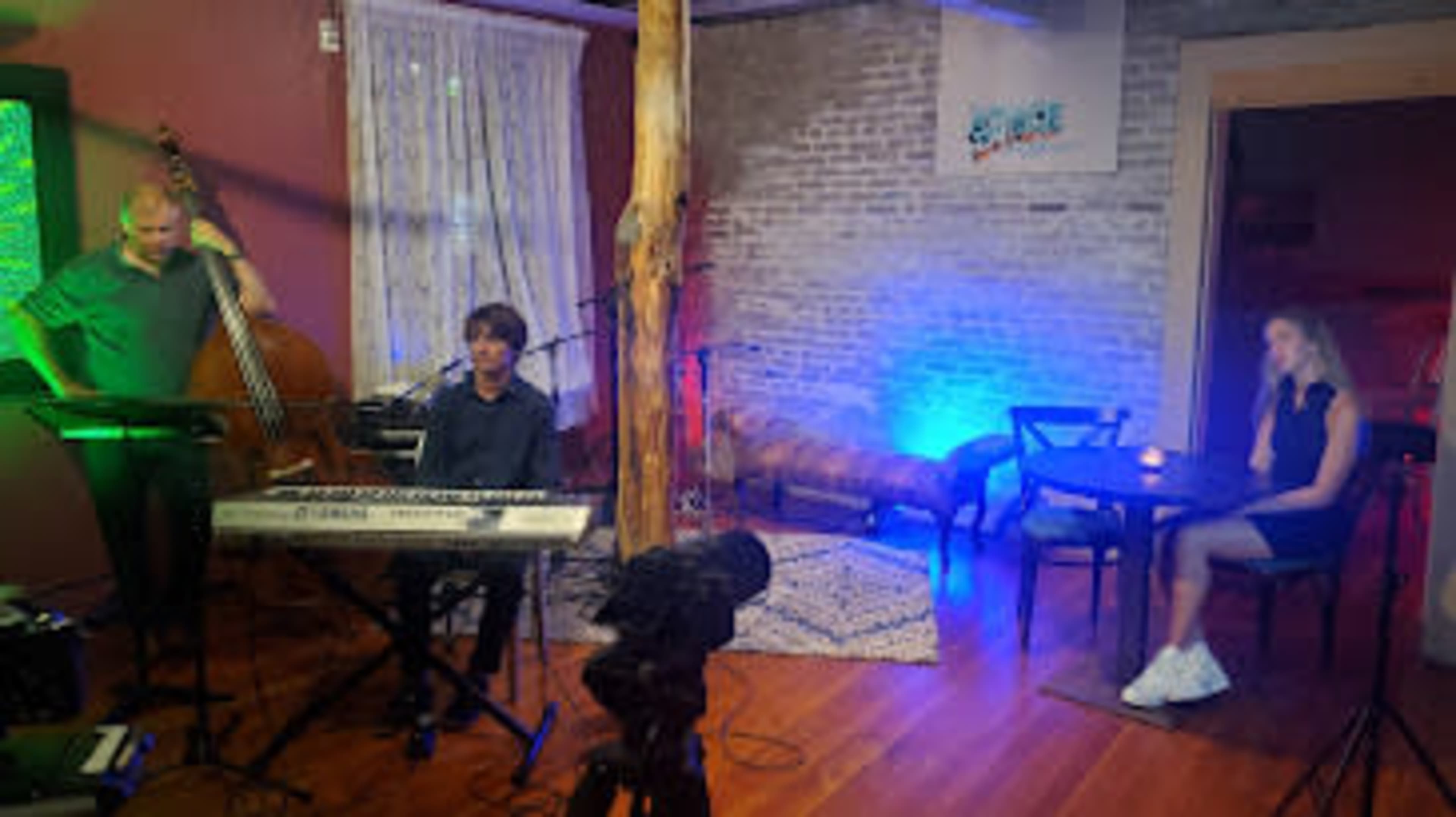 A musician plays a keyboard while a bassist stands nearby, and a woman sits at a table in a room with a rustic ambiance and soft lighting.