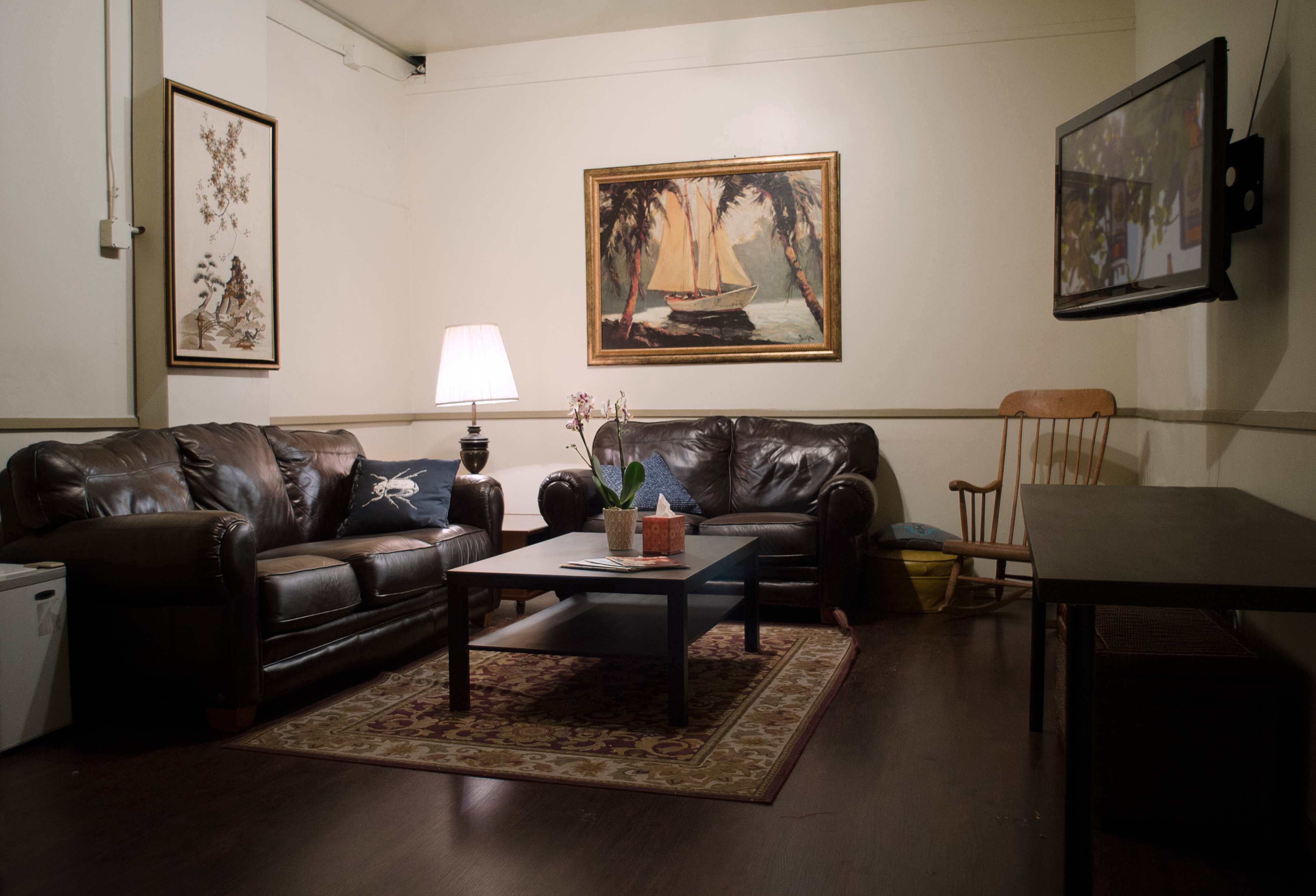 A living room features two leather sofas, a coffee table, a lamp, a rocking chair, and a television on the wall, with artwork depicting a sailboat.