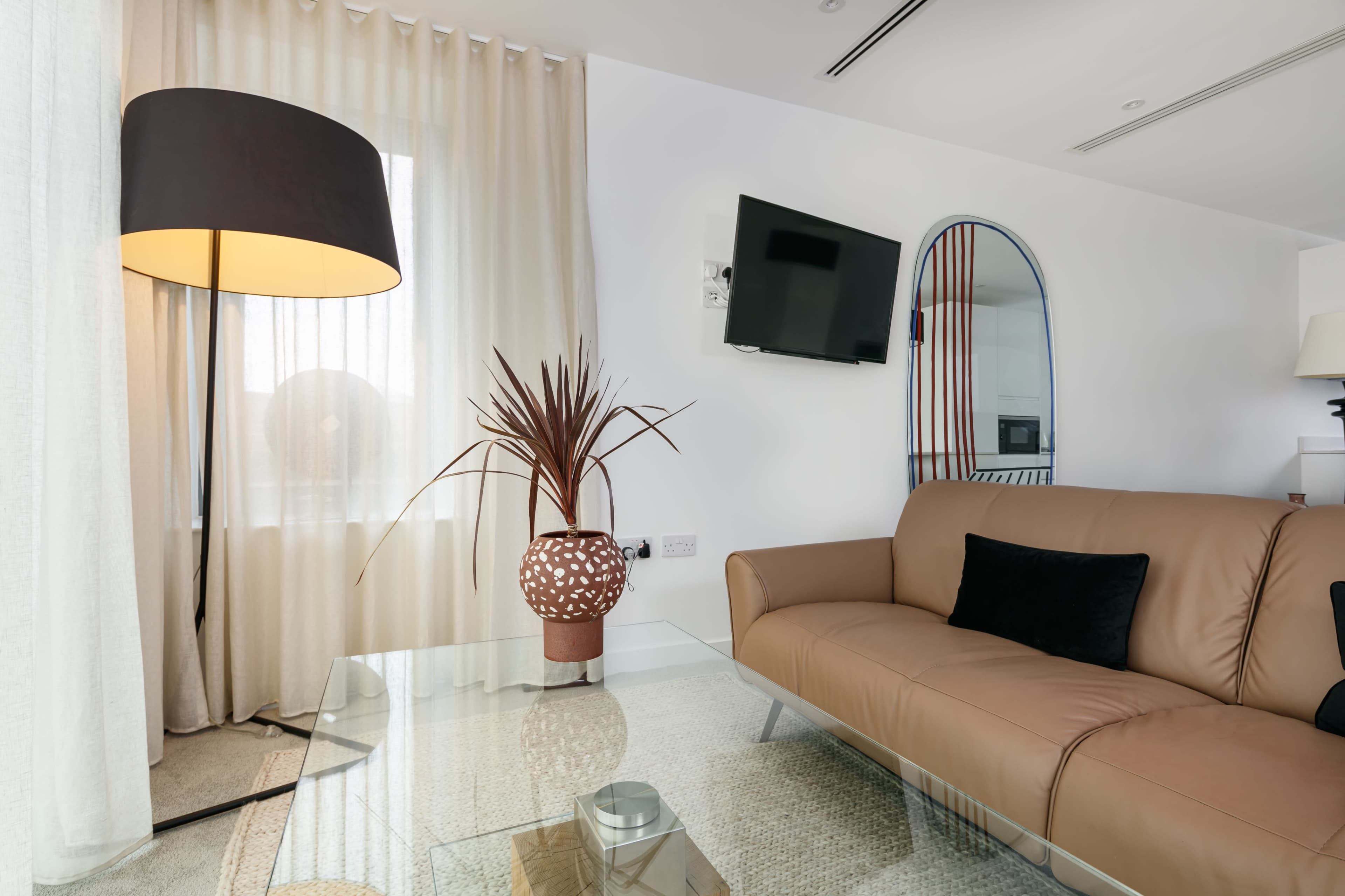 A modern living room features a leather sofa, a glass coffee table, a tall lamp, and a decorative plant beside sheer curtains and a wall-mounted TV.