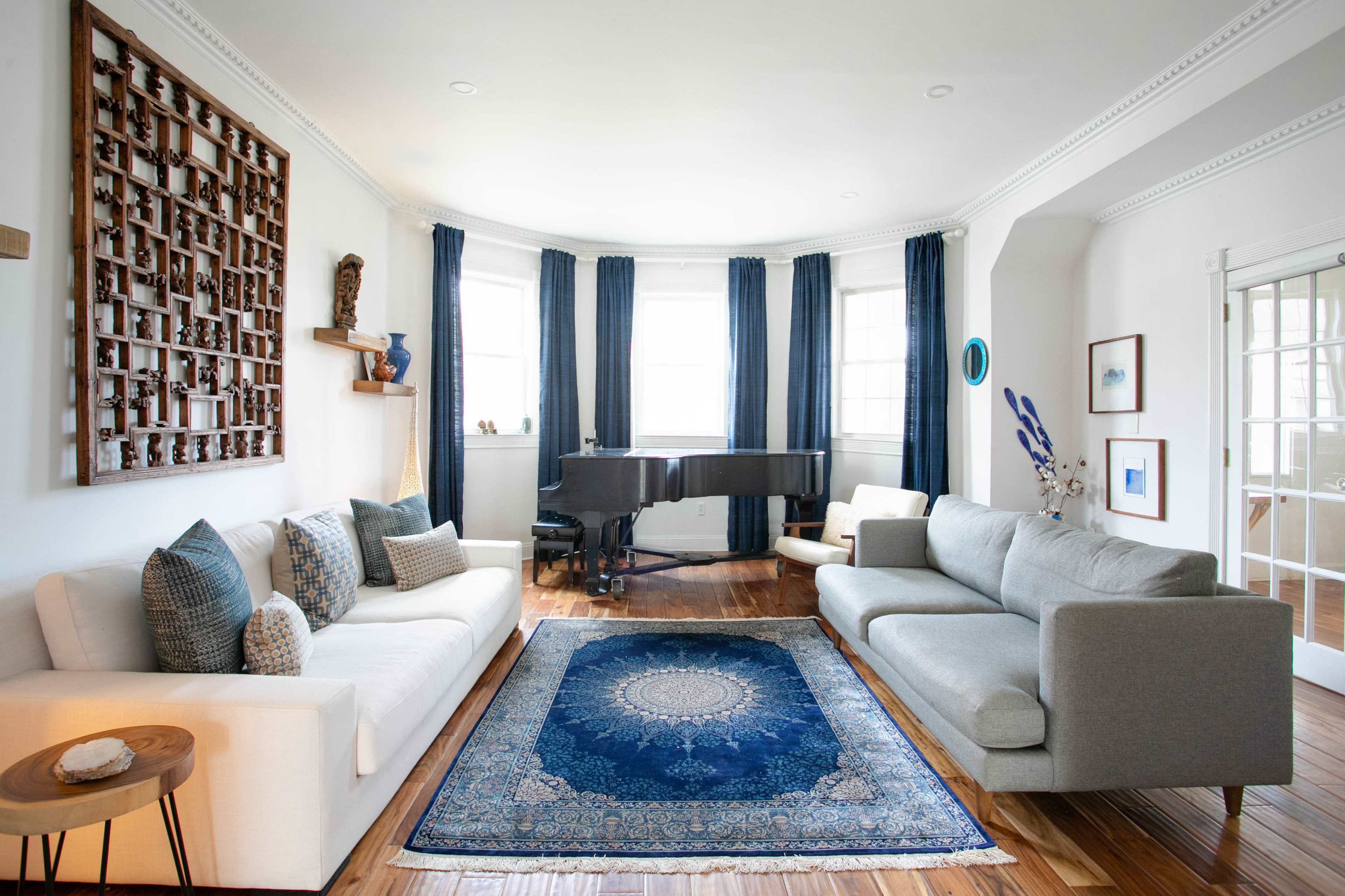 A bright living room featuring two sofas, a decorative wall display, a piano, and a blue patterned rug on wooden flooring.