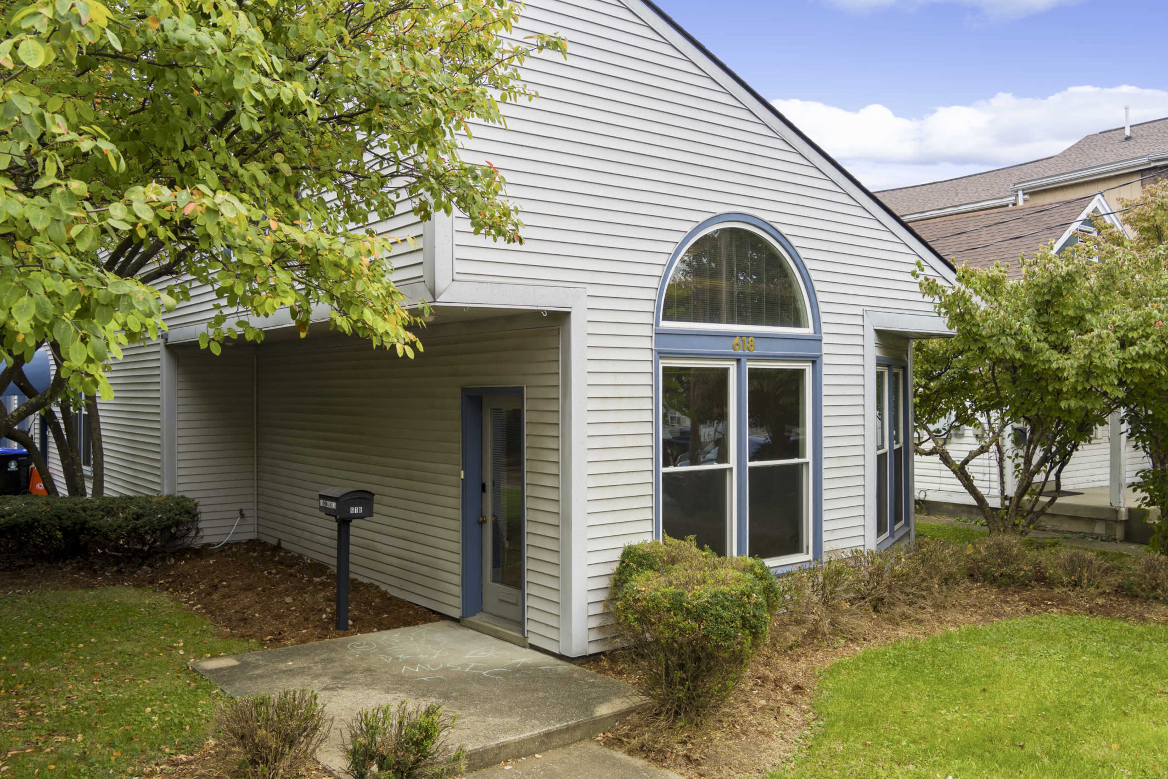 A single-story house with a peaked roof, large front windows, and a mailbox at the entrance, surrounded by shrubs and trees.