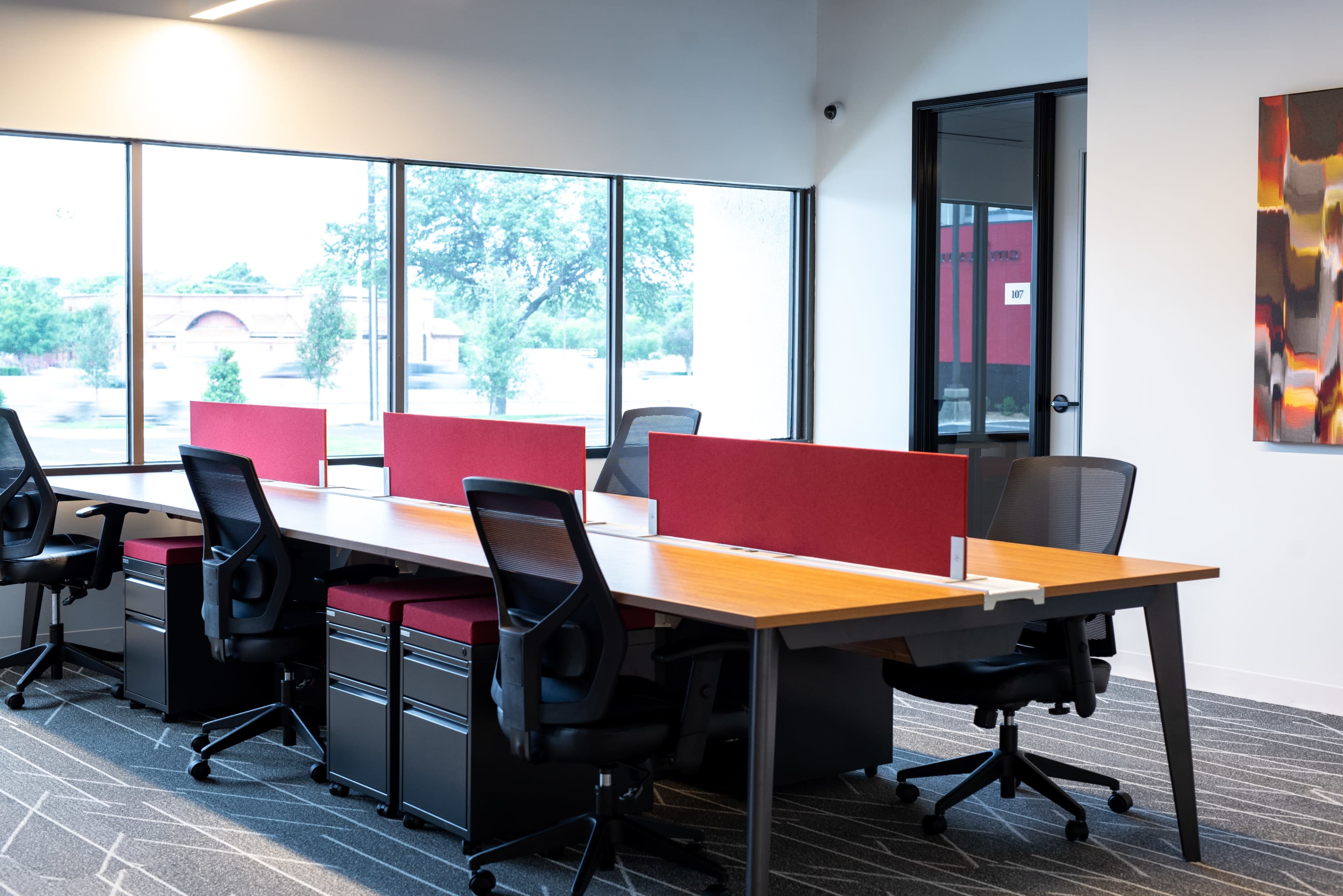 The image shows a modern office space with a long wooden table surrounded by black ergonomic chairs, partitioned by red panels, and large windows allowing natural light.