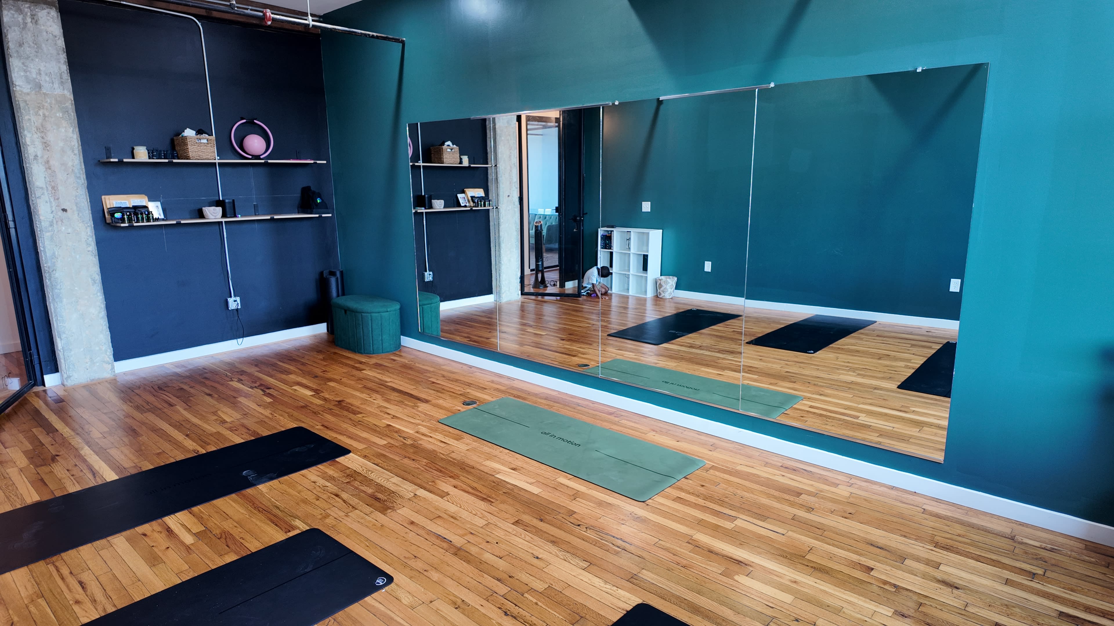 The image shows a yoga studio with wooden flooring, a green accent wall, and yoga mats laid out in front of a large mirror.