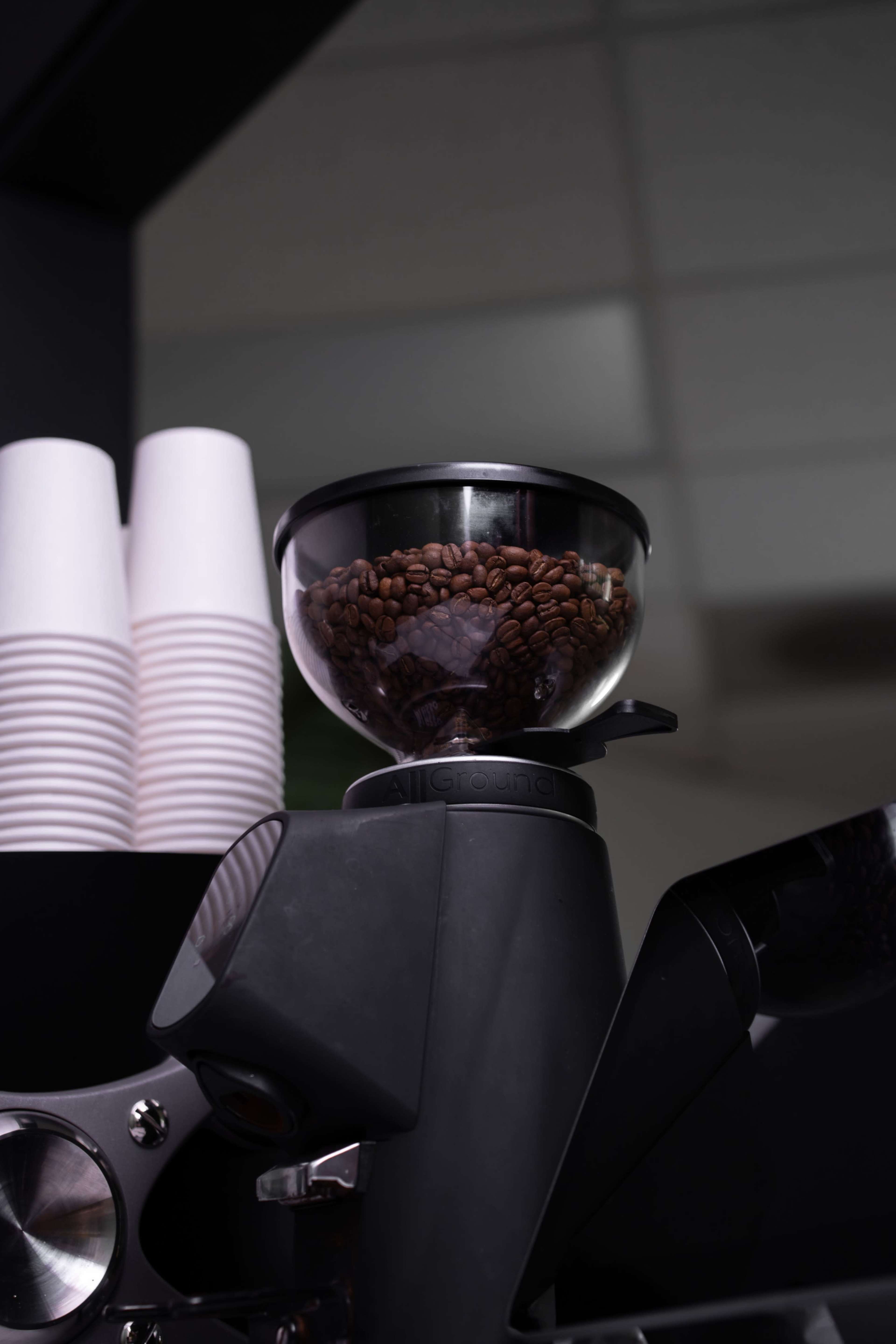 A coffee grinder with a glass hopper filled with coffee beans is positioned near a stack of disposable cups.