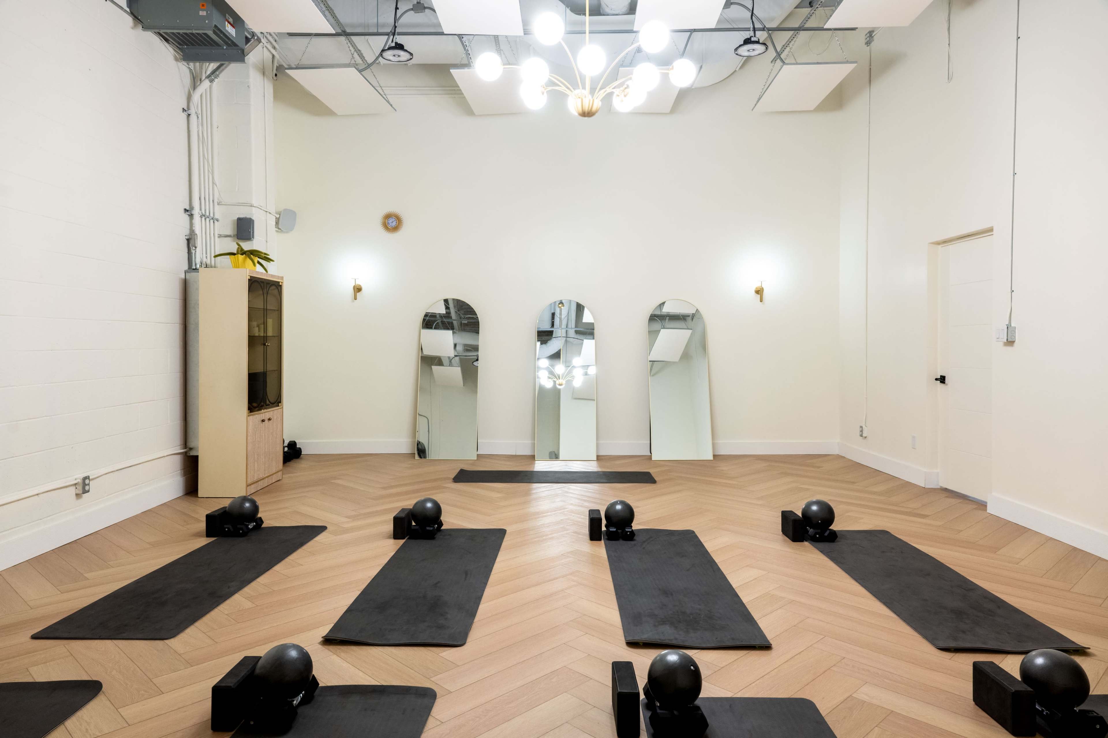 The image shows a minimalist exercise studio with several black yoga mats and weights arranged neatly on a light wood floor, complemented by mirrors and simple lighting fixtures.