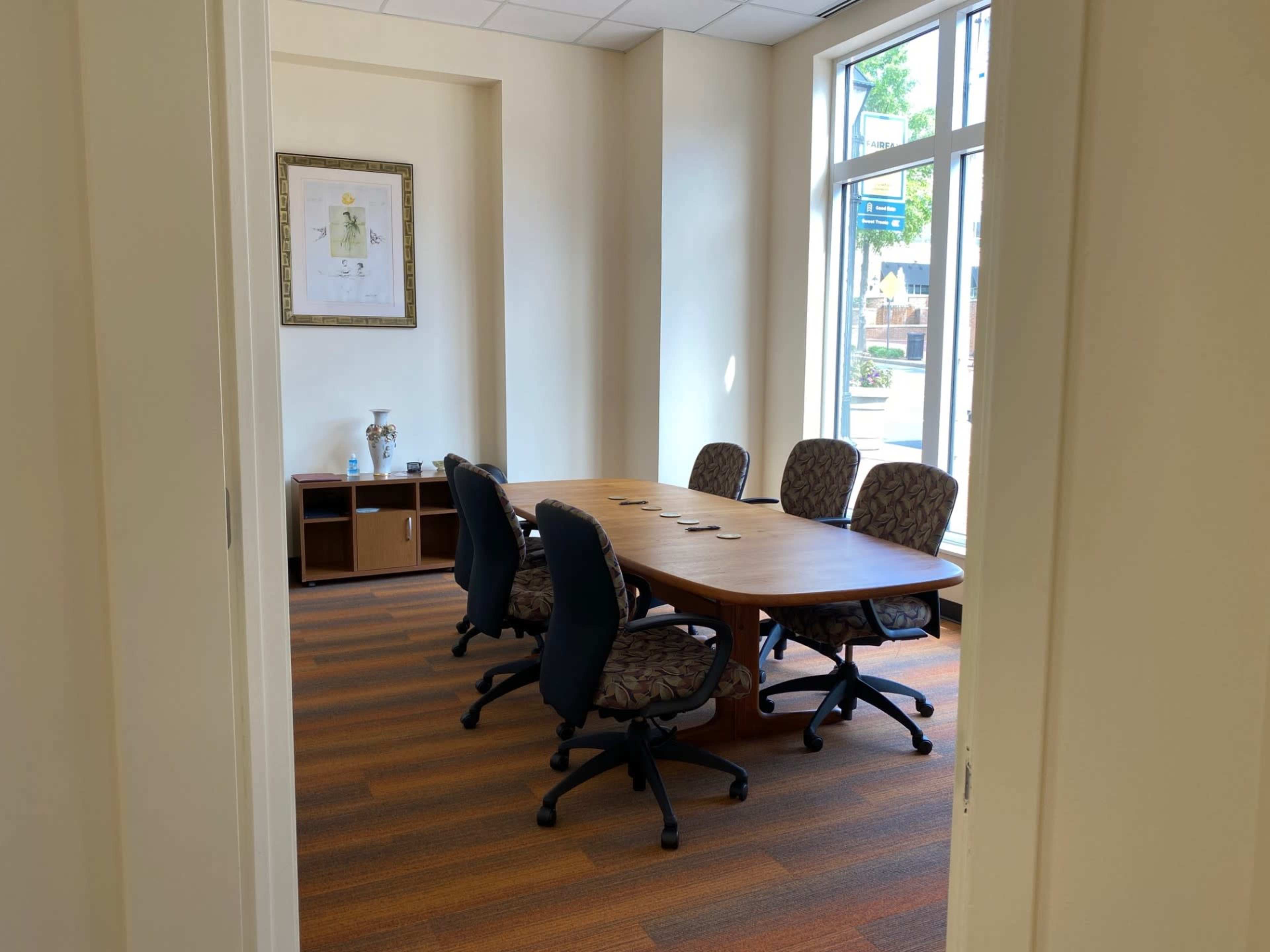 A vacant conference room features a large wooden table surrounded by five office chairs, with a framed artwork on the wall.