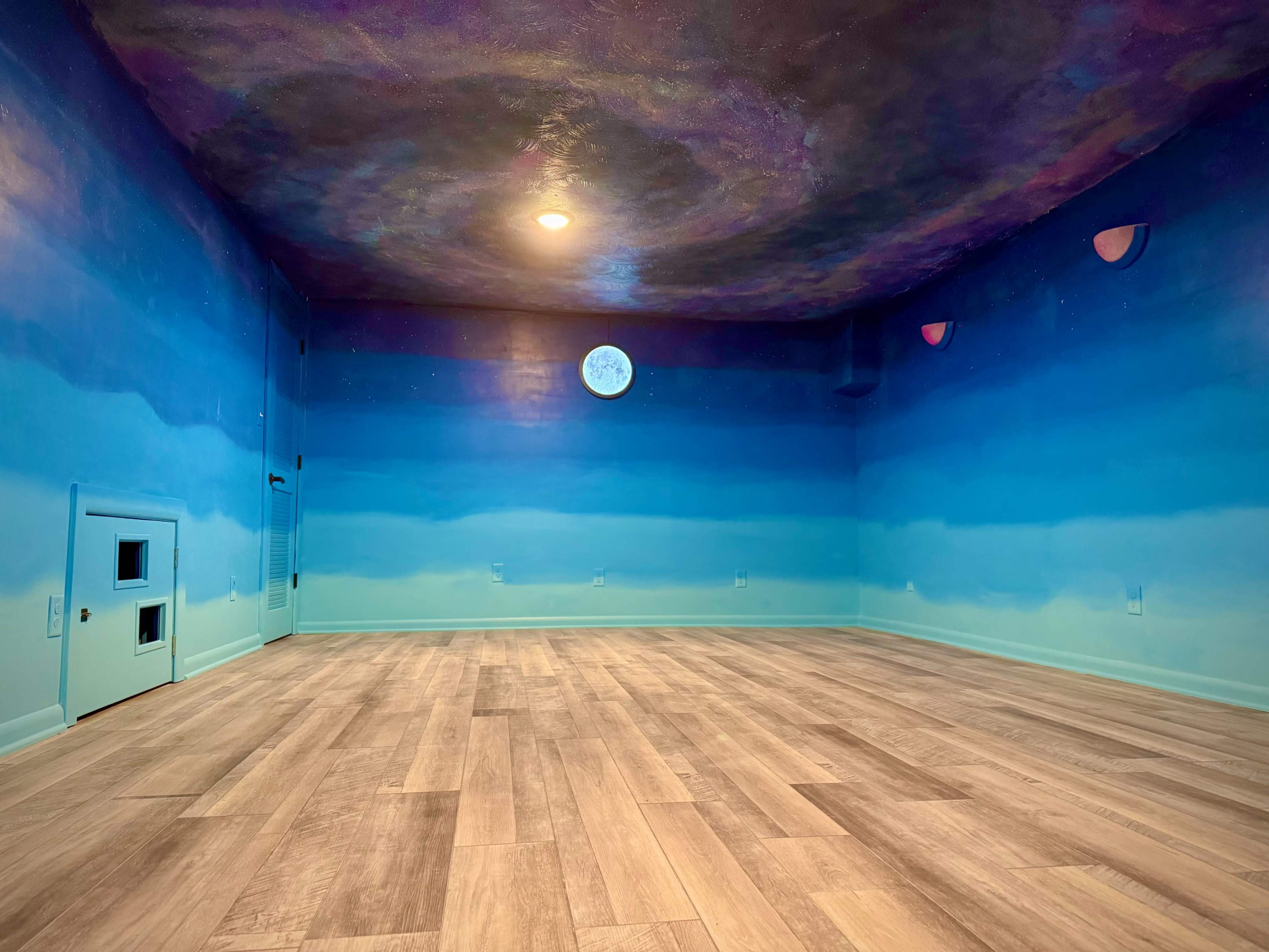 The image shows a room with a painted blue gradient mural on the walls and ceiling, featuring a moon and soft pink accents, with wooden flooring and no furniture inside.