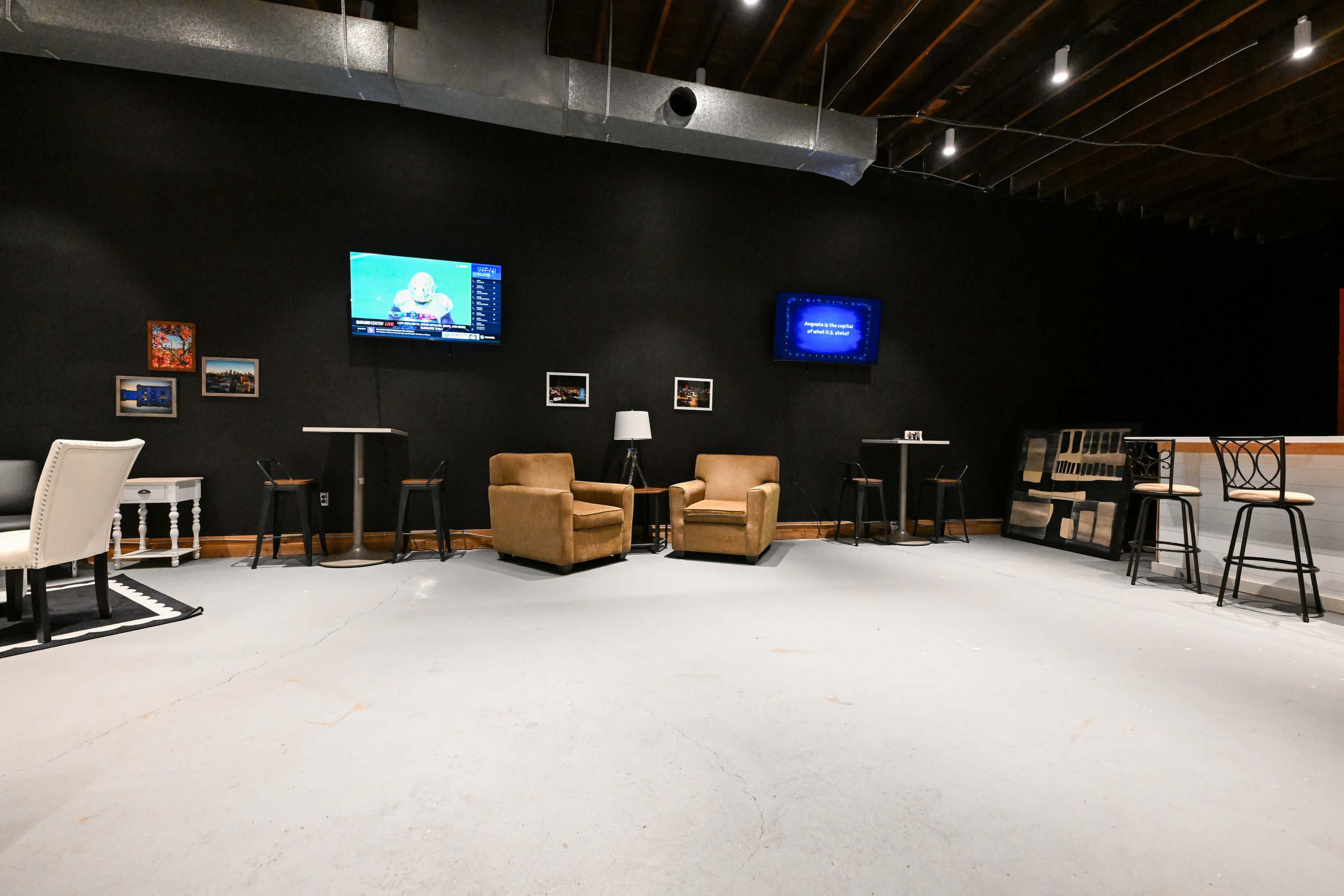 The image shows a spacious room with a concrete floor, featuring two brown armchairs, several small tables, two televisions mounted on the wall, and a counter along one side.