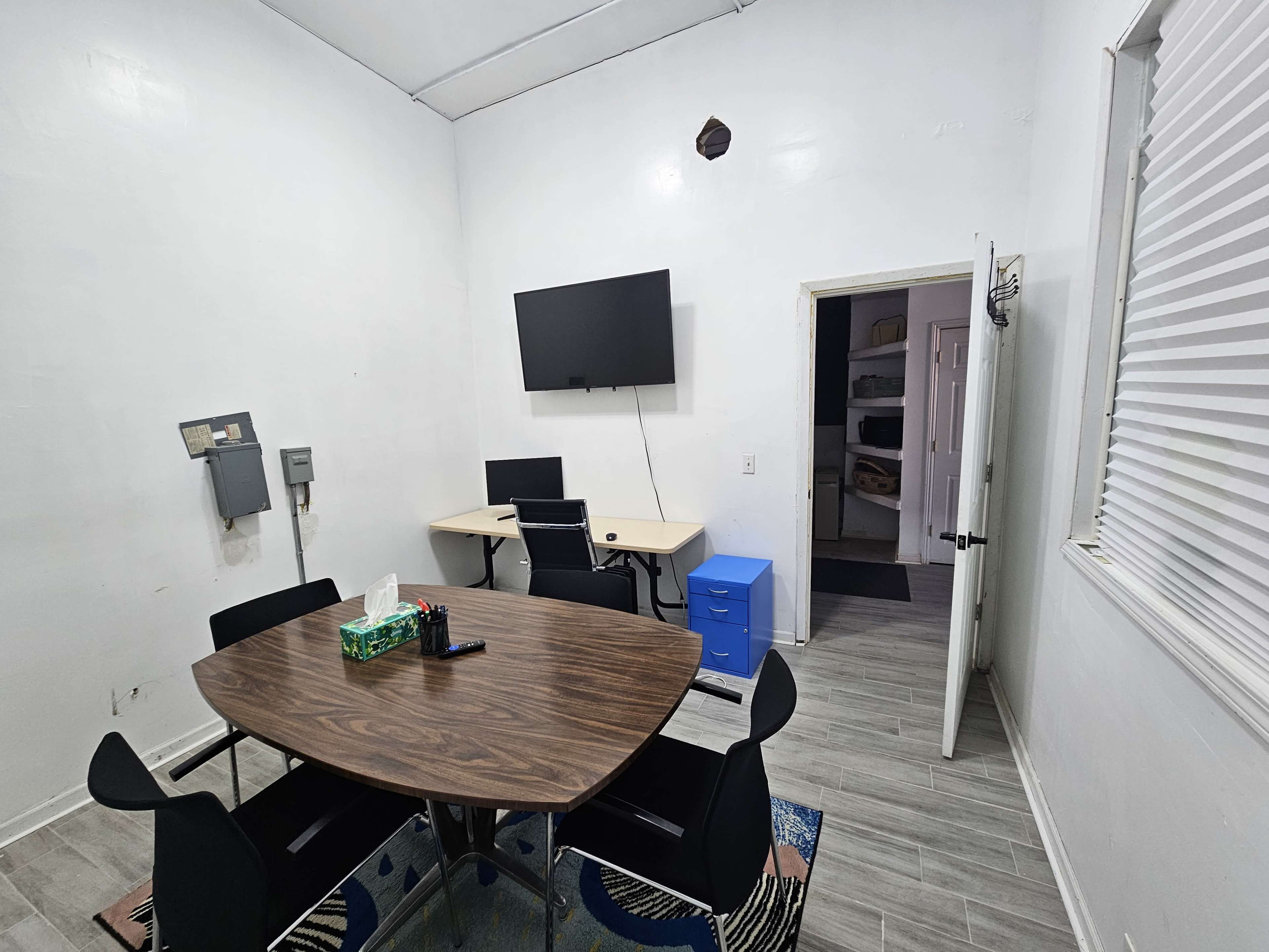 The image shows a small, bright room with a round wooden table surrounded by black chairs, a television mounted on the wall, and a desk with a blue filing cabinet visible in the background.