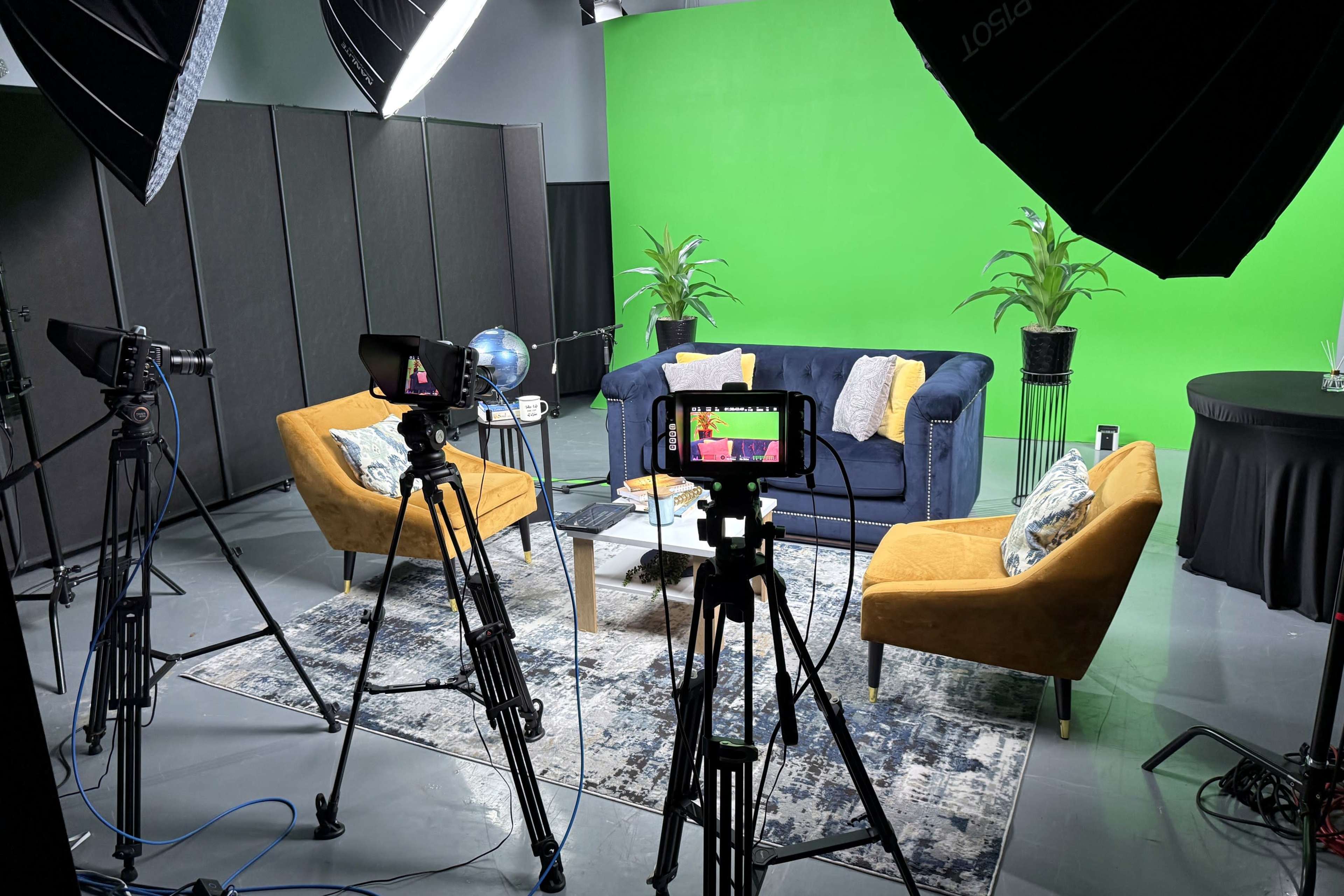 The image shows a studio setup with two colorful chairs, a table, a globe, and camera equipment positioned around a green screen backdrop.