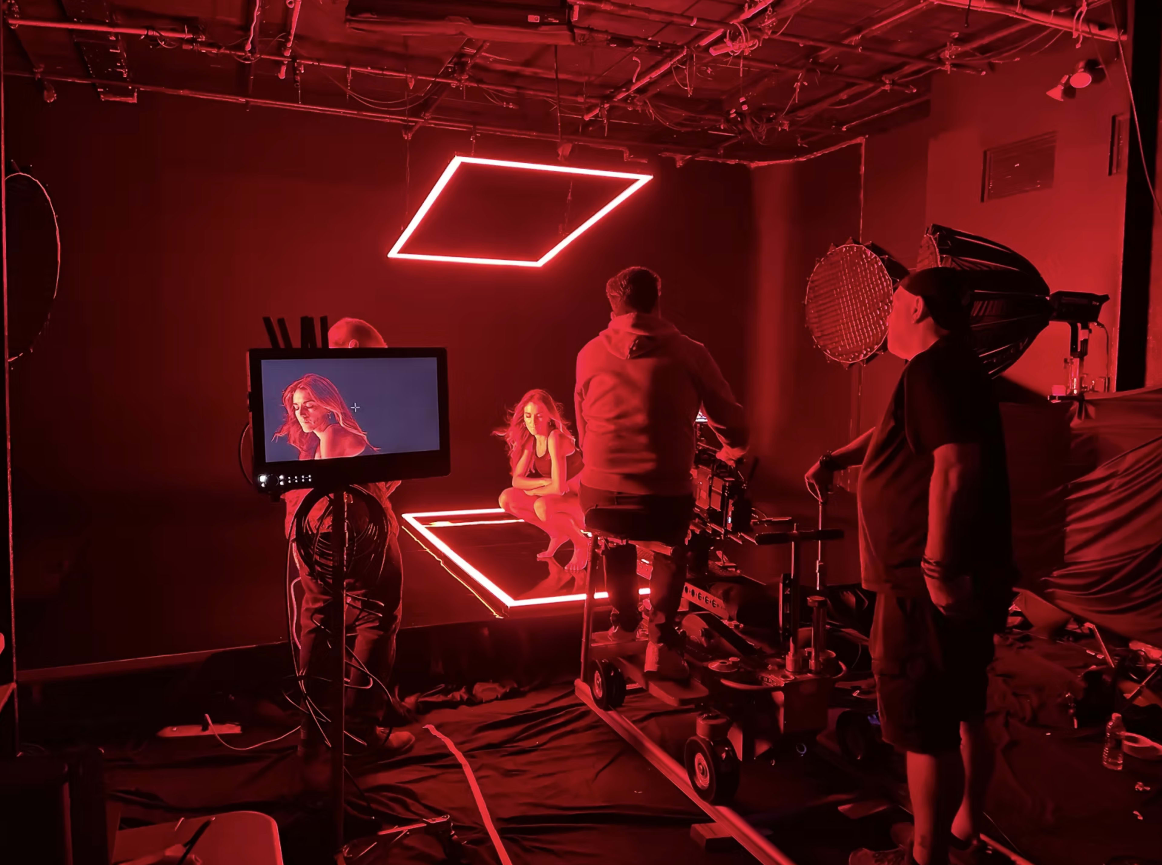 A film crew is captured working on a set featuring red neon lighting, with a camera focused on a model posing amid the illuminated backdrop.