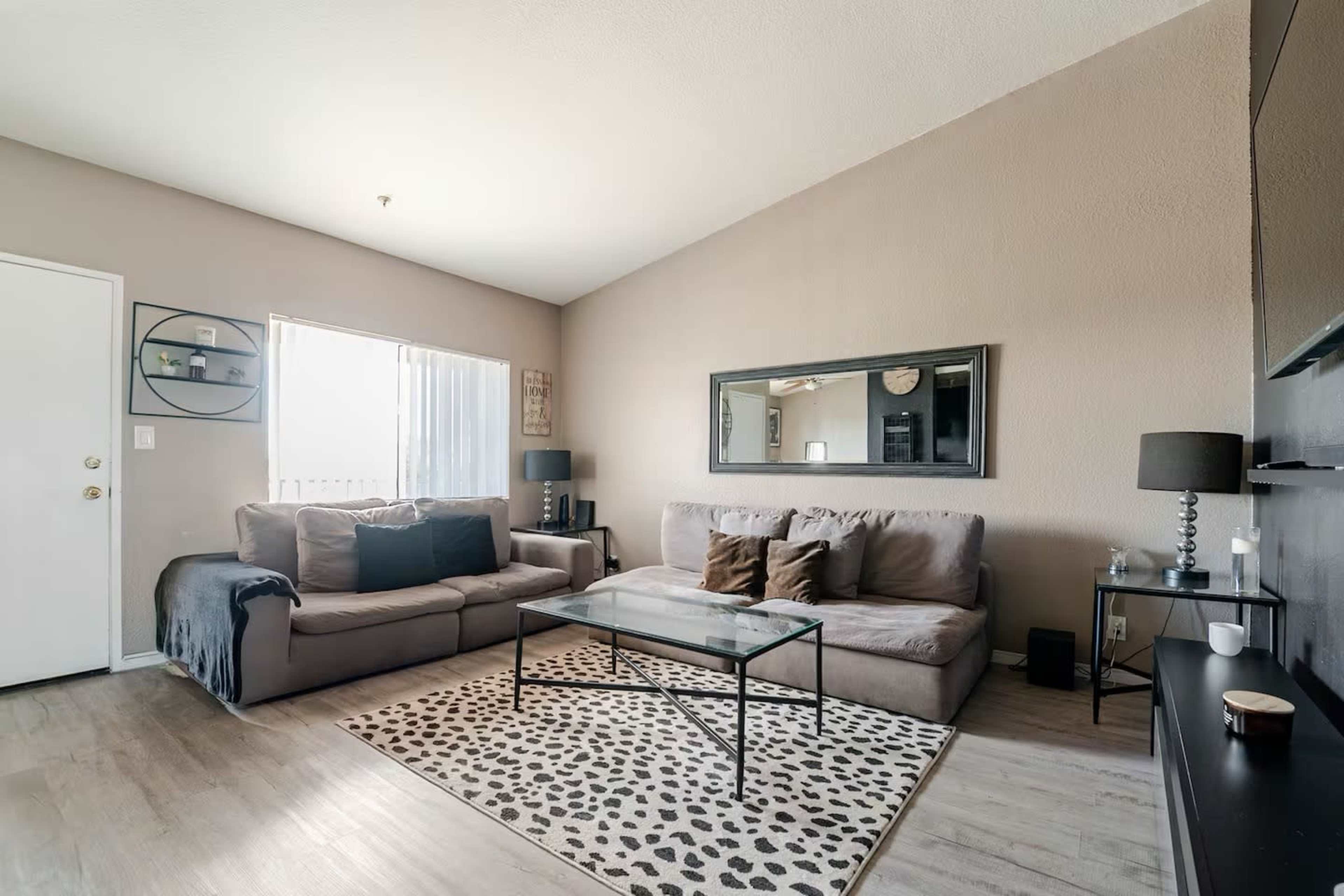 The image shows a living room with two gray sofas, a glass coffee table on a leopard-print rug, and a large wall mirror.