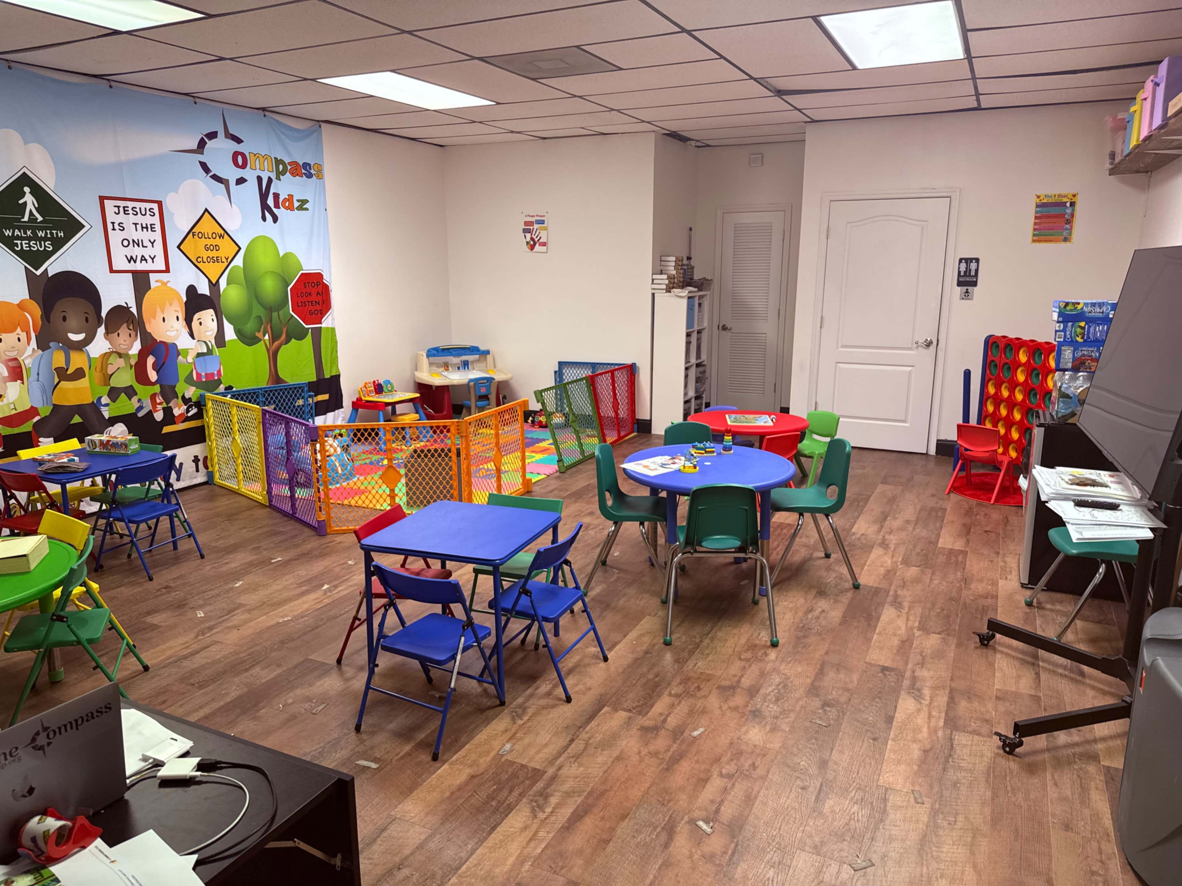 The image shows a brightly colored children’s playroom with various tables and chairs, a playpen area, and a wall mural featuring cartoon characters.