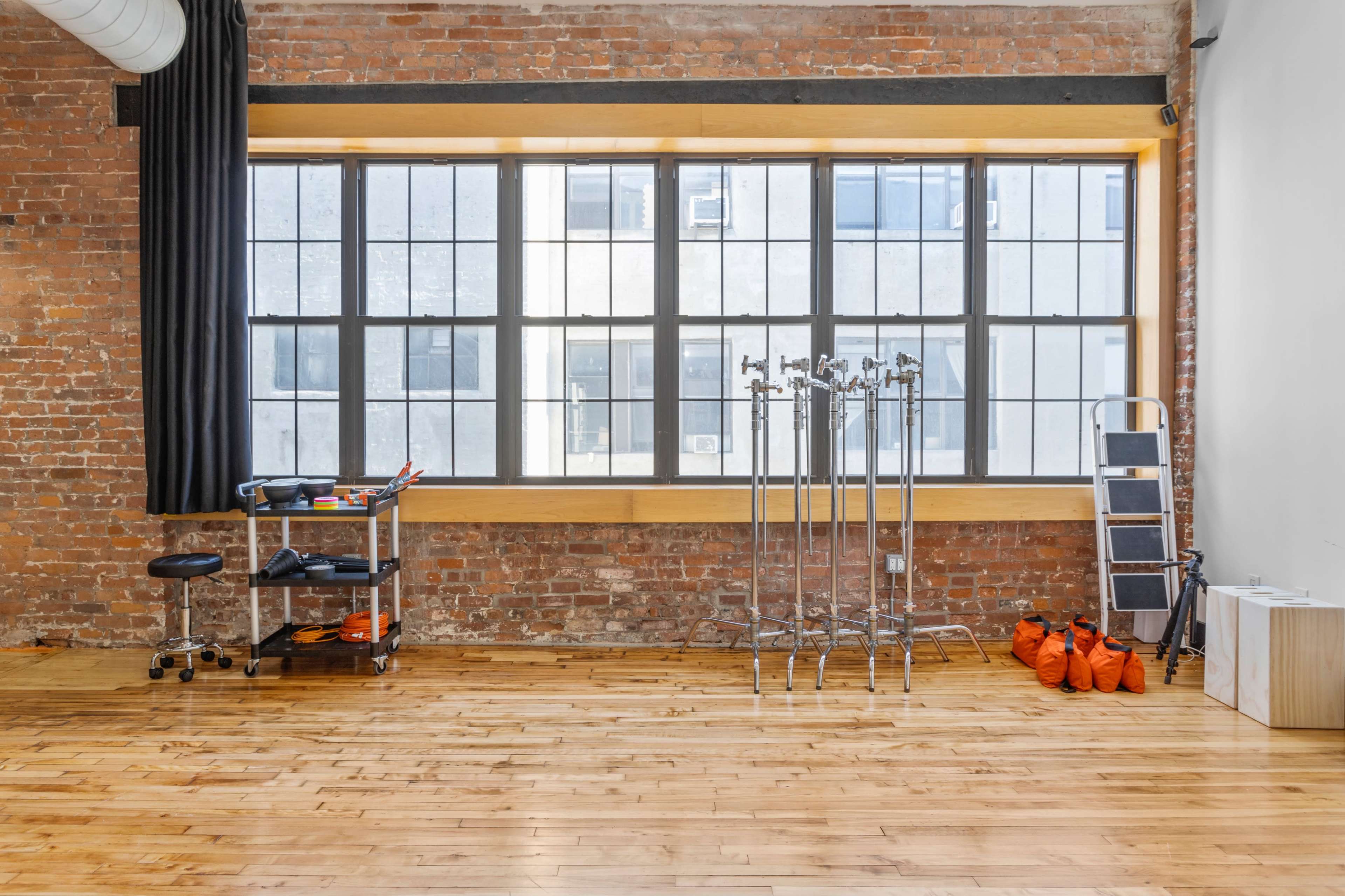 A spacious, minimalist room with exposed brick walls, a large window, and equipment including a cart and stands along one side.