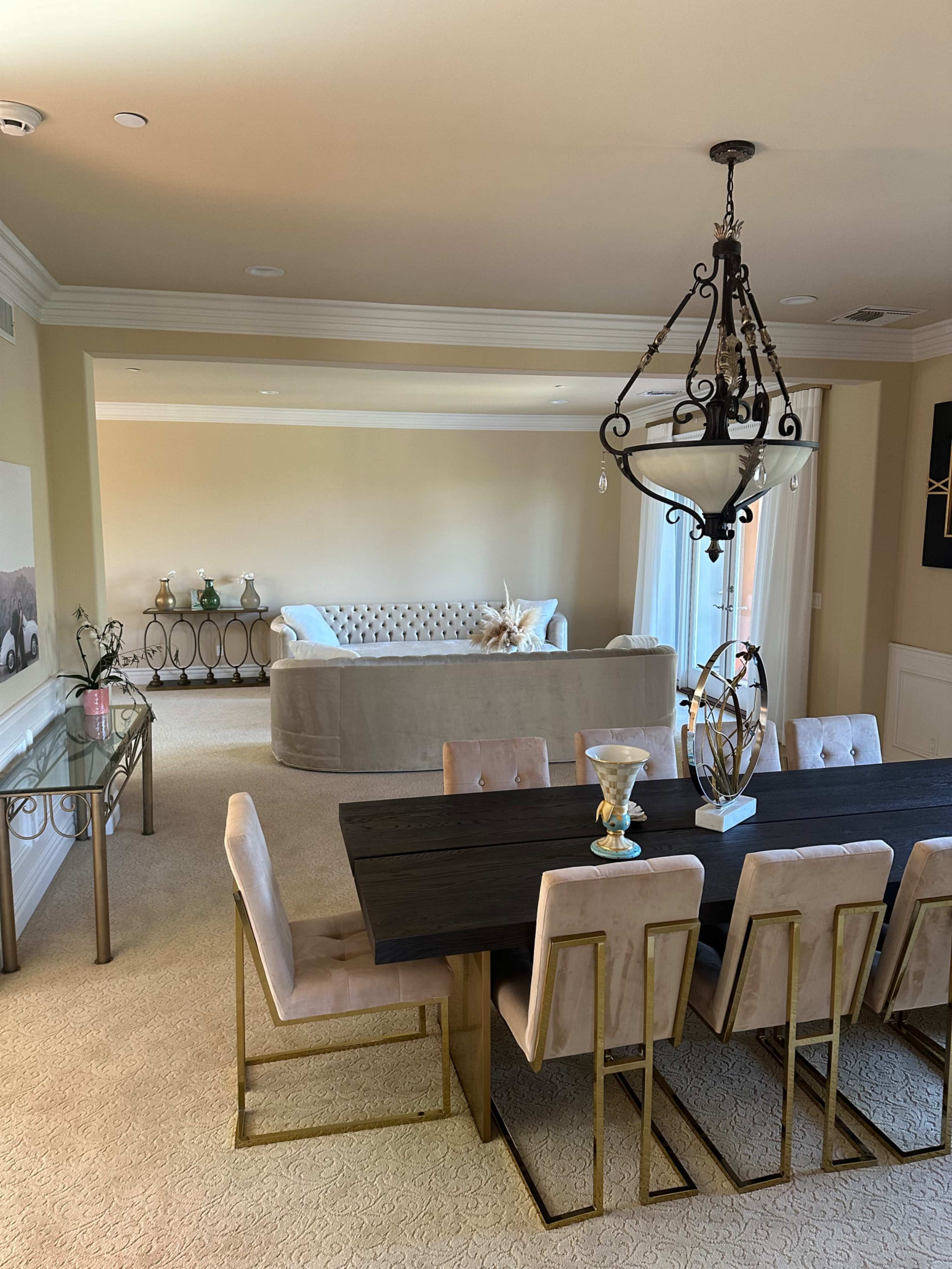 A dining area features a long black table with gold-accented chairs, adjacent to a spacious living room with a light-colored sofa.