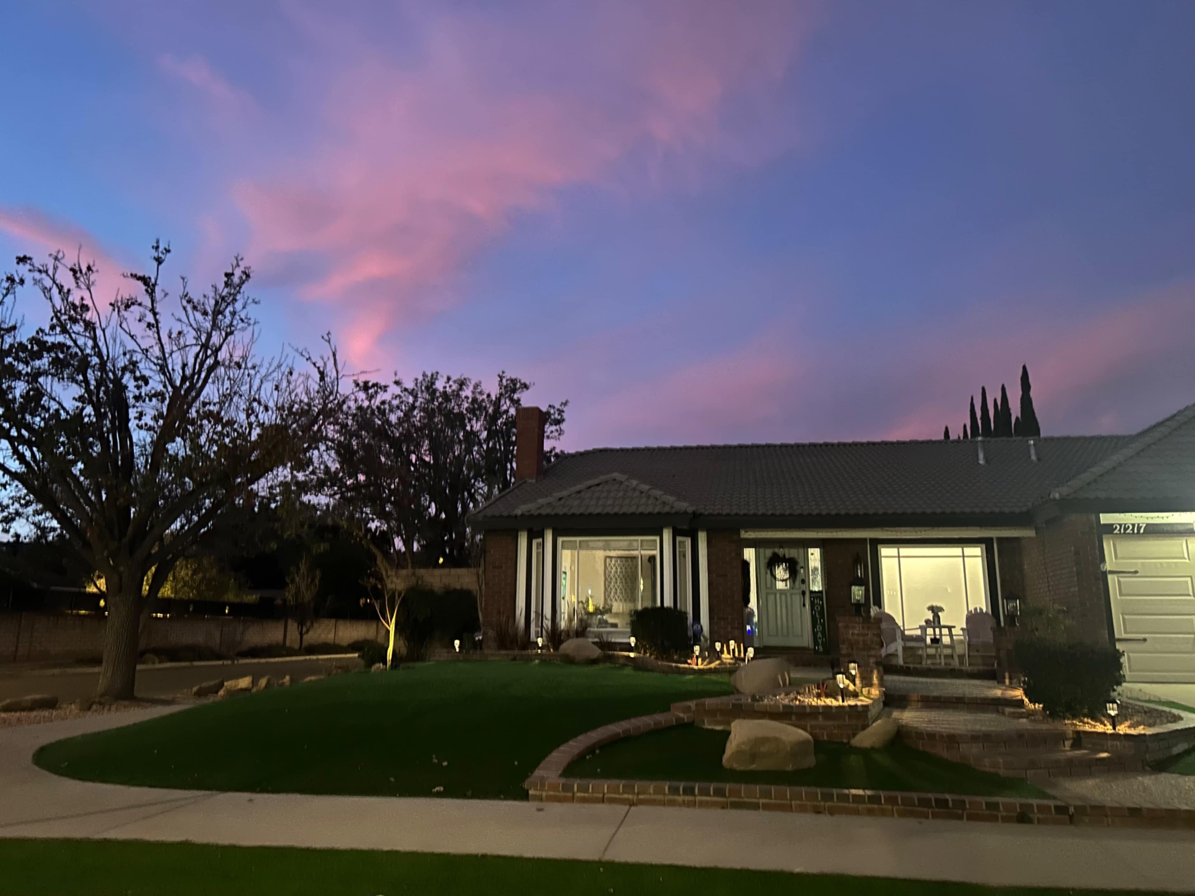 A single-story brick house with a landscaped front yard is set against a colorful sunset sky.