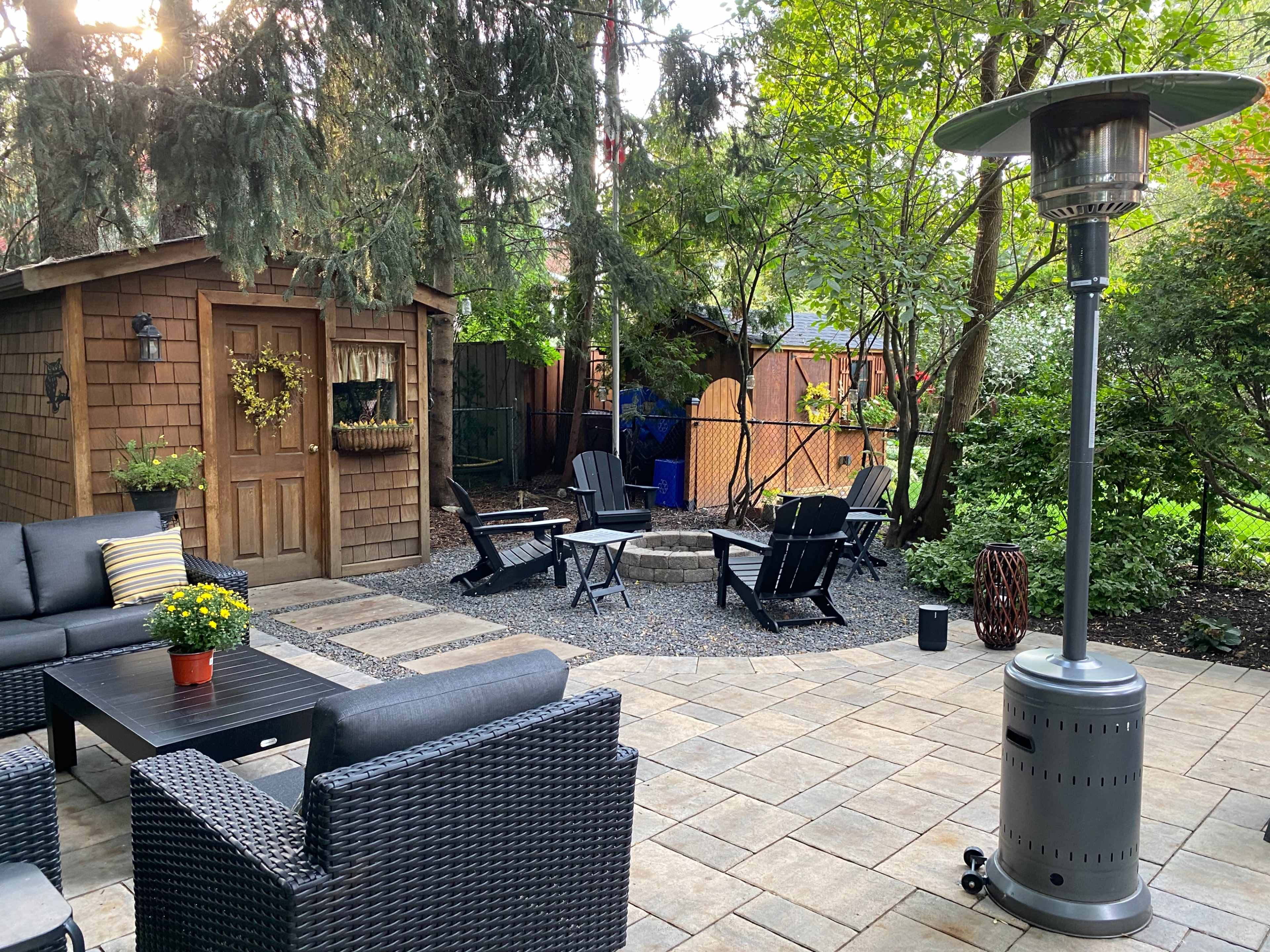 A cozy outdoor patio features a seating area with black furniture, a fire pit, and a patio heater surrounded by trees and a shed.