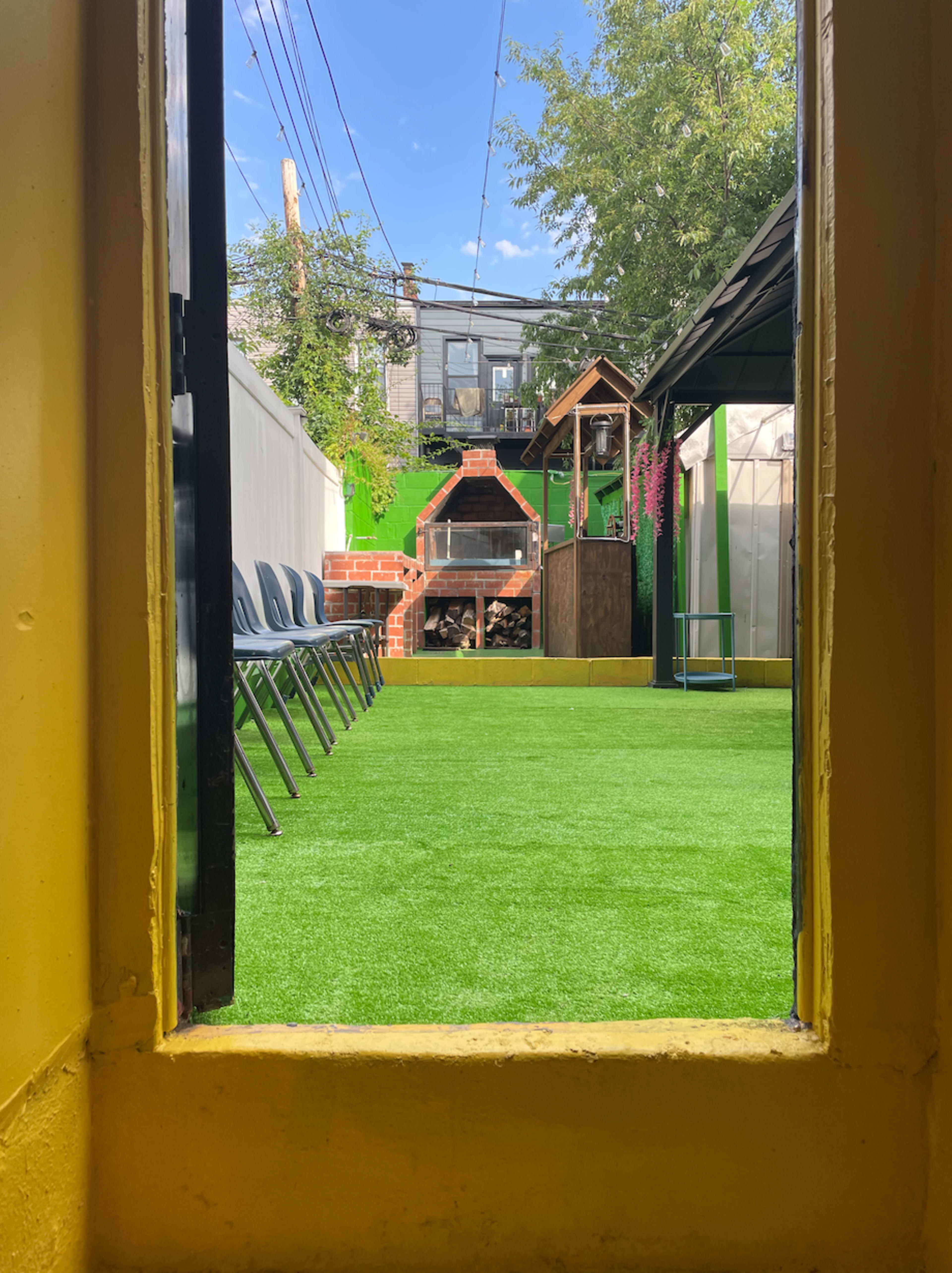 A view through a doorway shows a green artificial lawn area with a barbecue pit, wooden storage, and seating chairs arranged along one side.