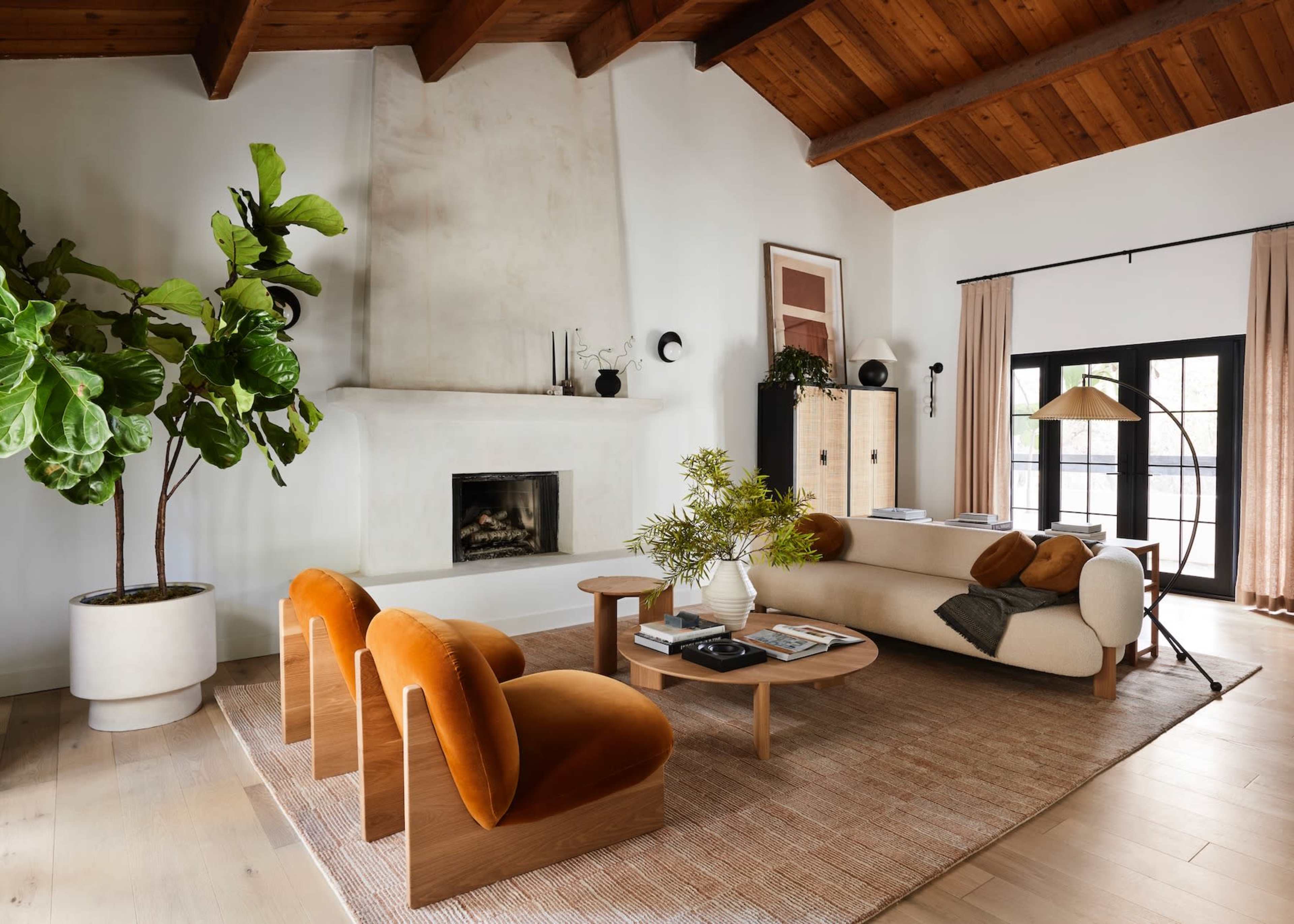 The living room features a high wooden ceiling, a white stone fireplace, a neutral sofa with orange accent chairs, and a large indoor plant.