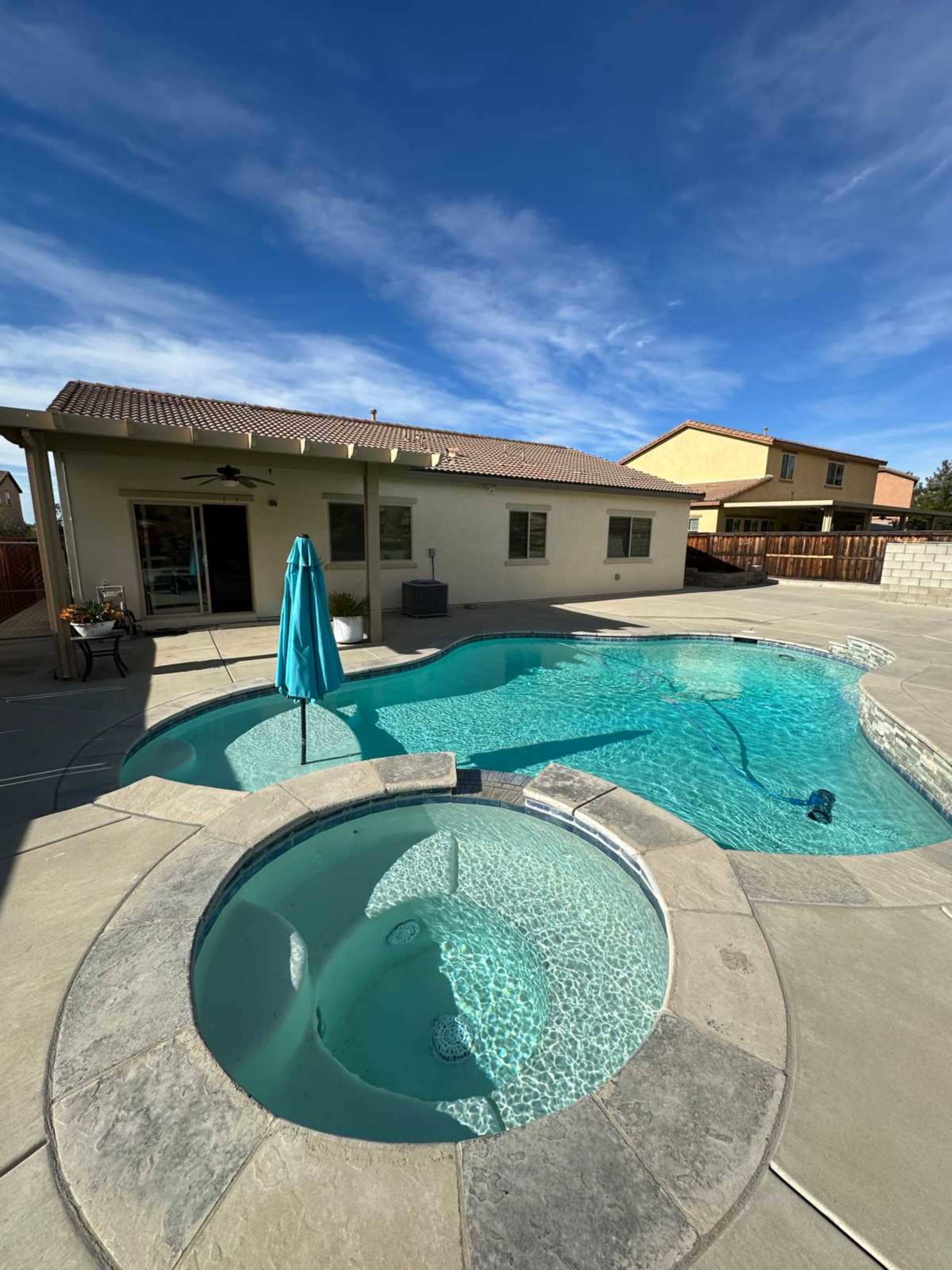 The image shows a backyard with a swimming pool and a spa, surrounded by concrete and a few homes in the background under a clear blue sky.