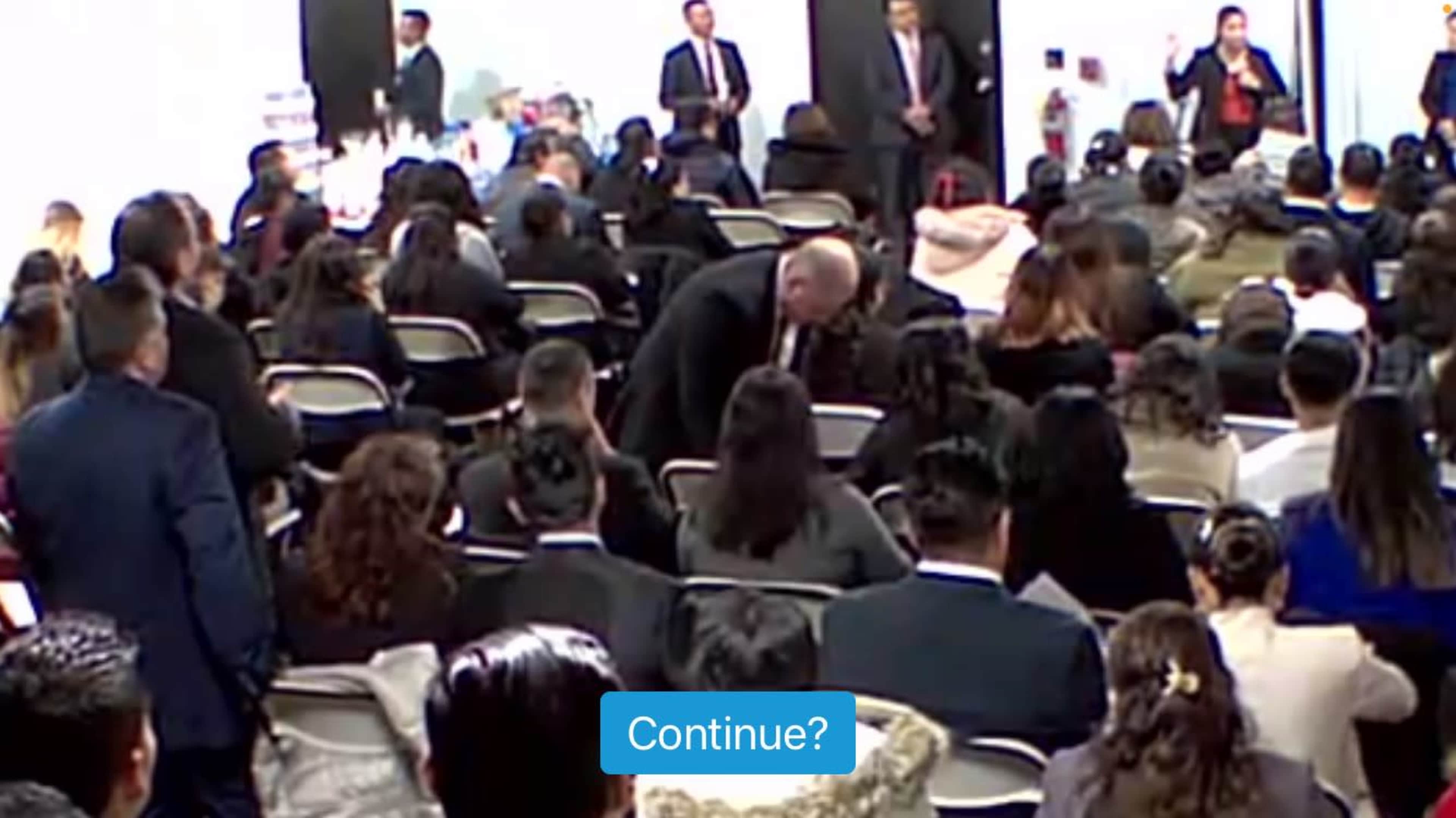 A large crowd of people is seated in a hall, with someone speaking at the front and a few individuals standing nearby.