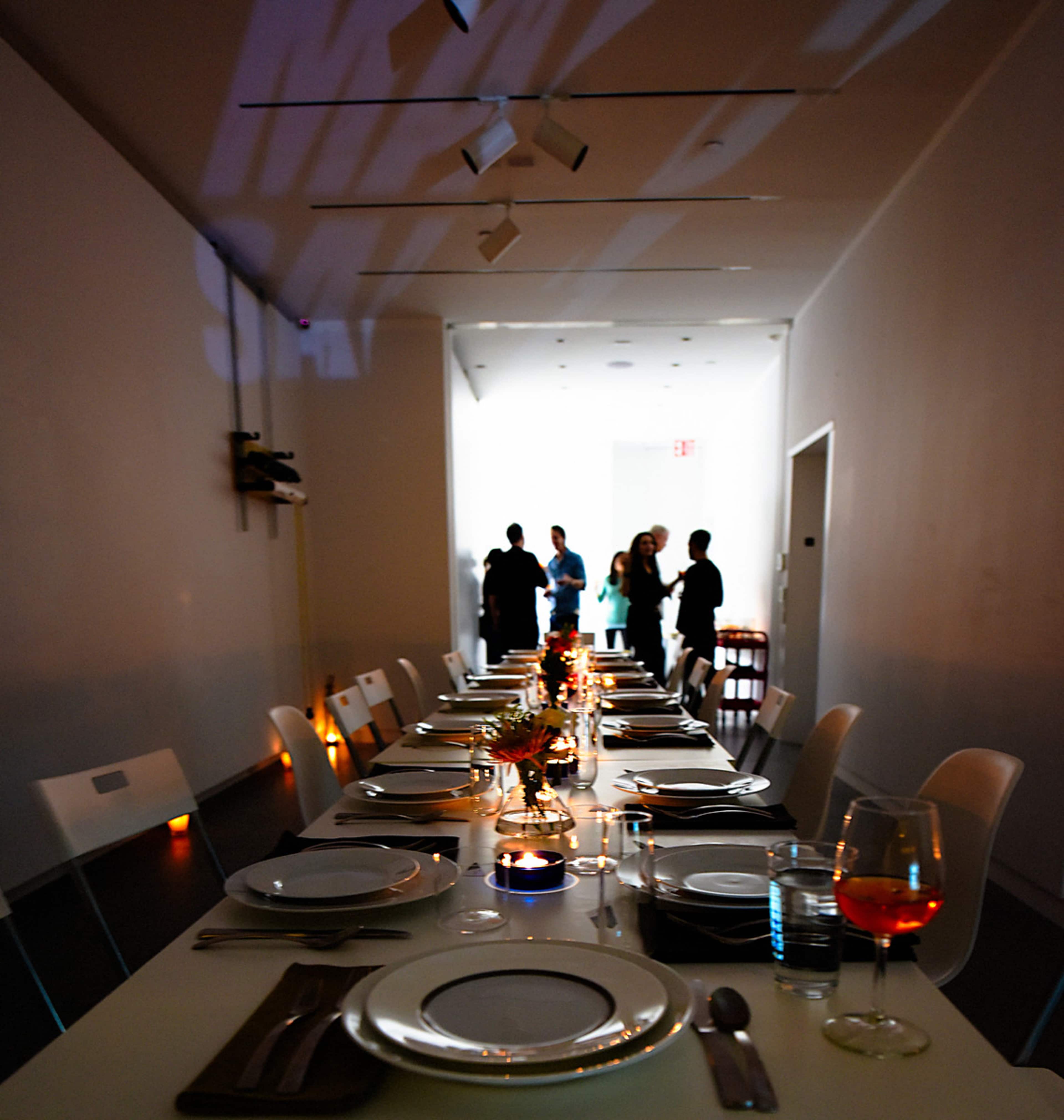 A long dining table is elegantly set with plates and glassware, while people gather in the background of a well-lit room.
