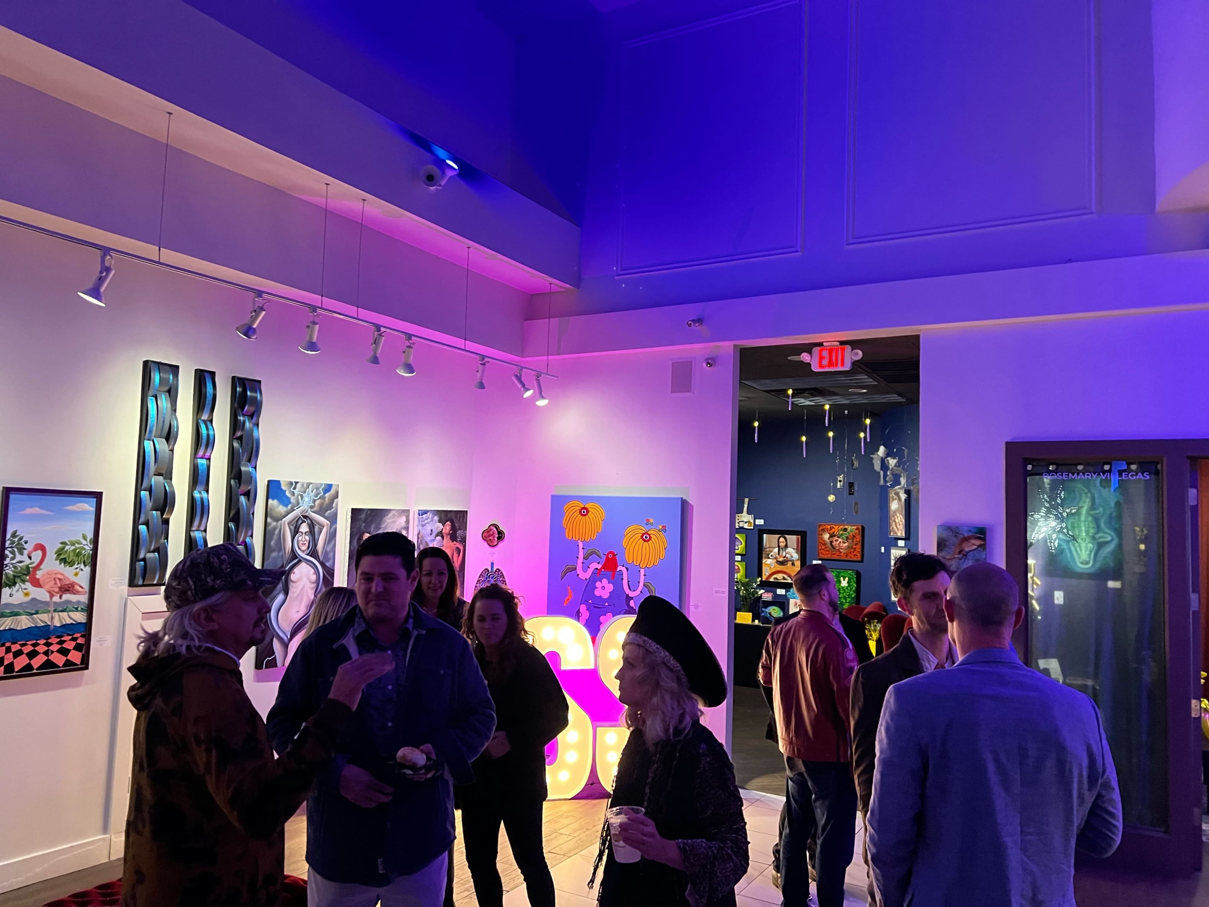 People socialize during an art exhibition featuring colorful artwork and vibrant lighting in a gallery space.