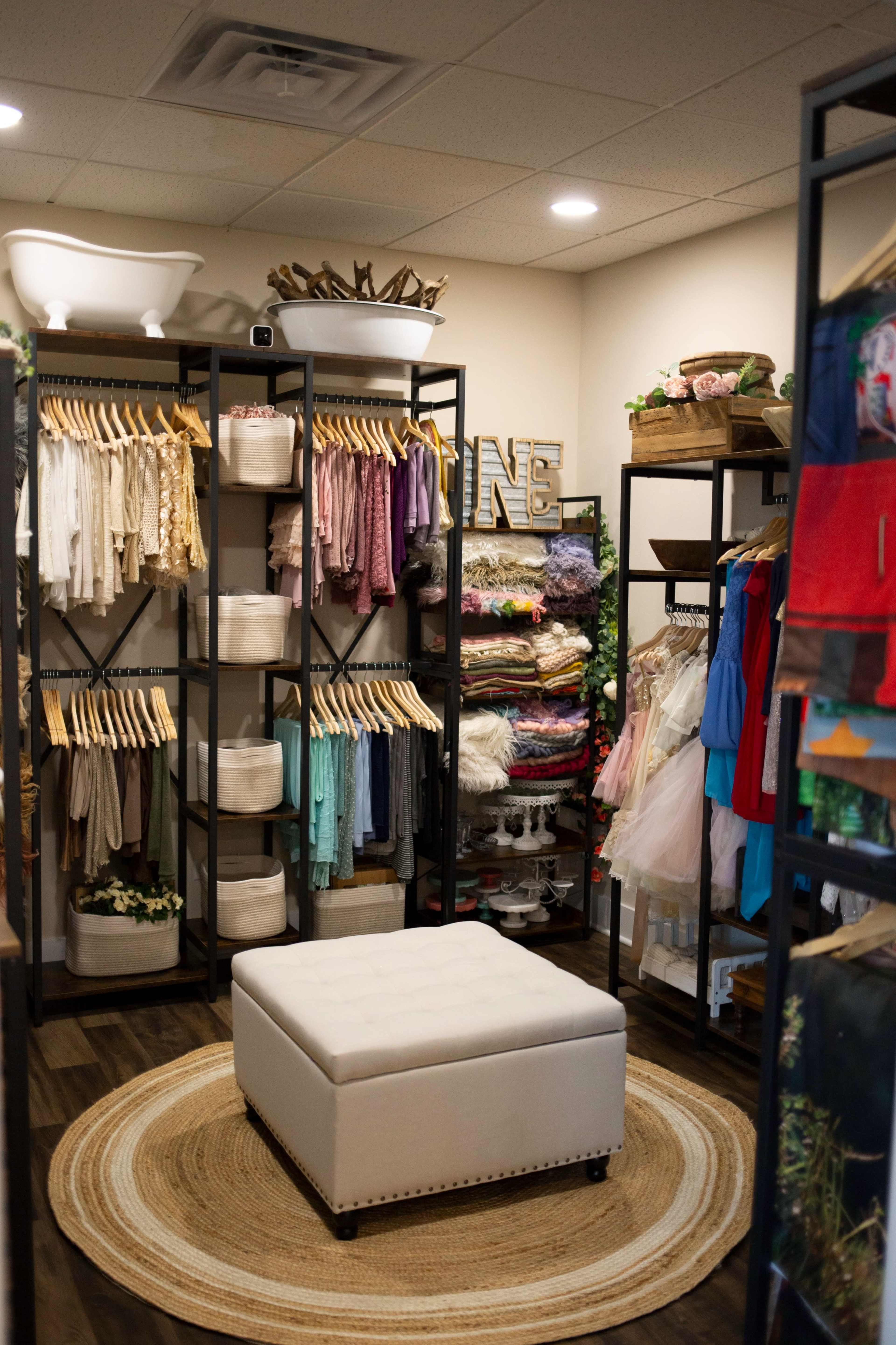 The image shows a neatly organized boutique featuring clothing racks, shelves with folded fabrics, and a central ottoman on a round rug.
