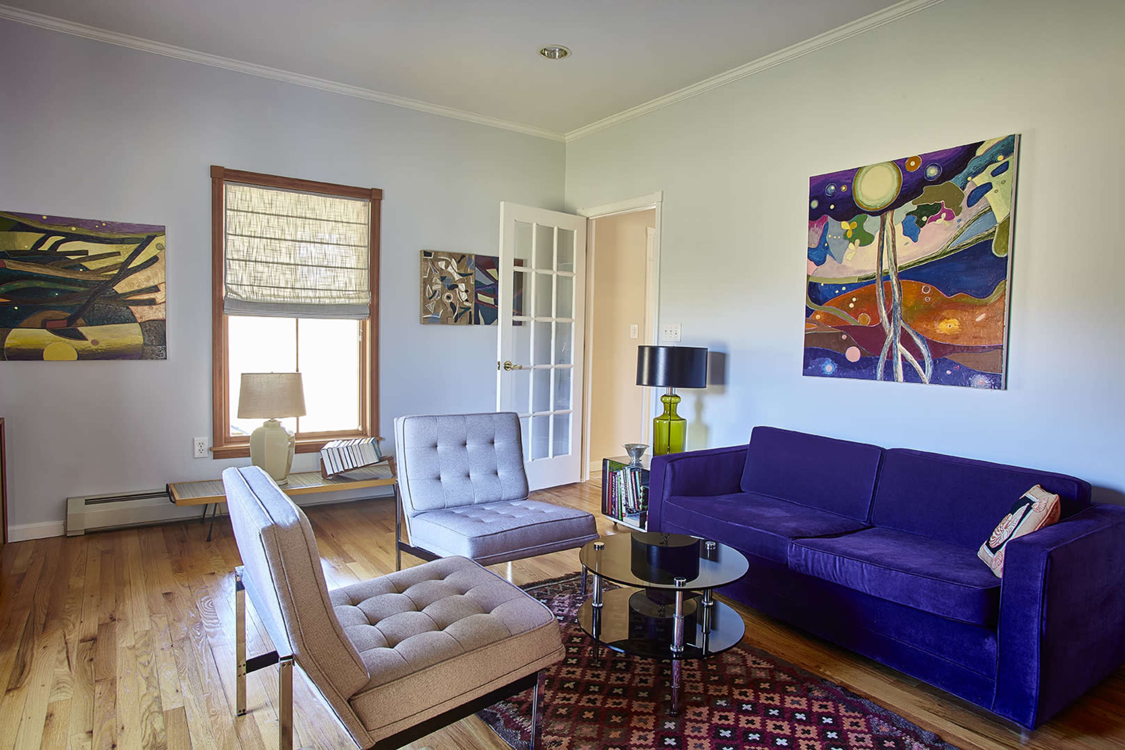 A living room with a purple sofa, two gray armchairs, a glass coffee table, and colorful artwork on the walls.