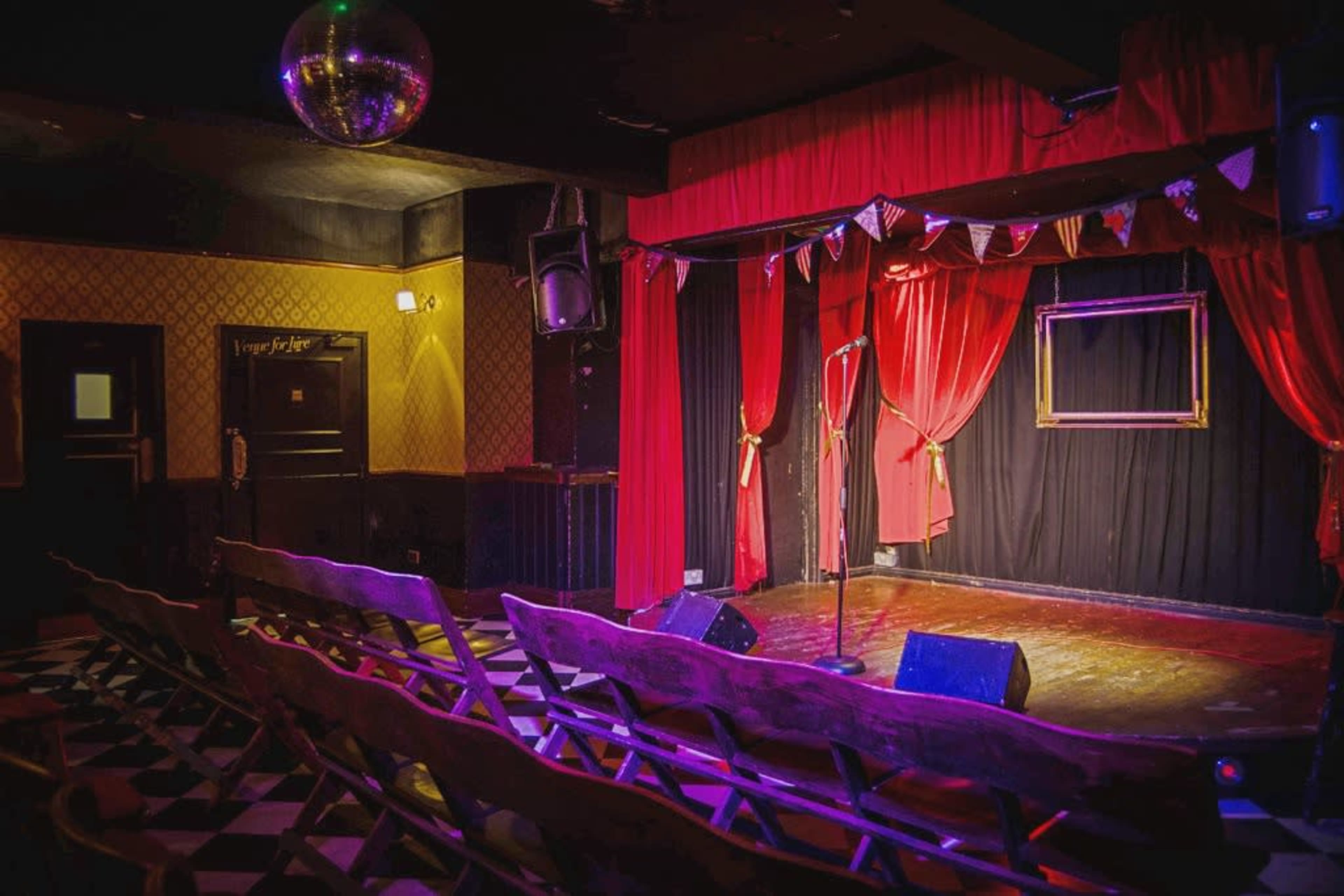 A small stage with a microphone stand, surrounded by red curtains and empty wooden benches facing the performance area, illuminated by colorful lights and a disco ball hanging from the ceiling.