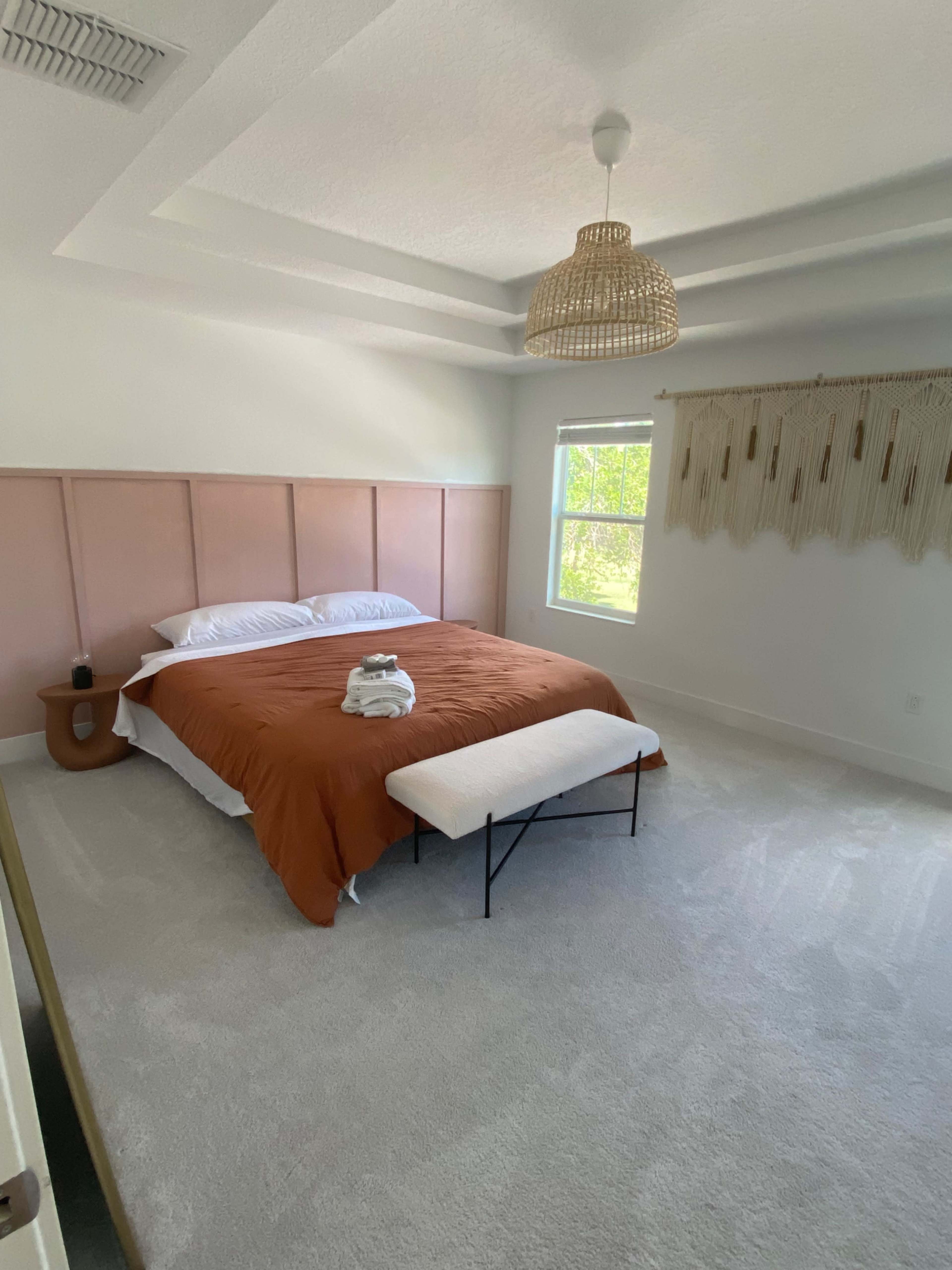 A spacious bedroom features a large bed with an orange duvet, a bench at the foot, and a decorative wall hanging above a window.