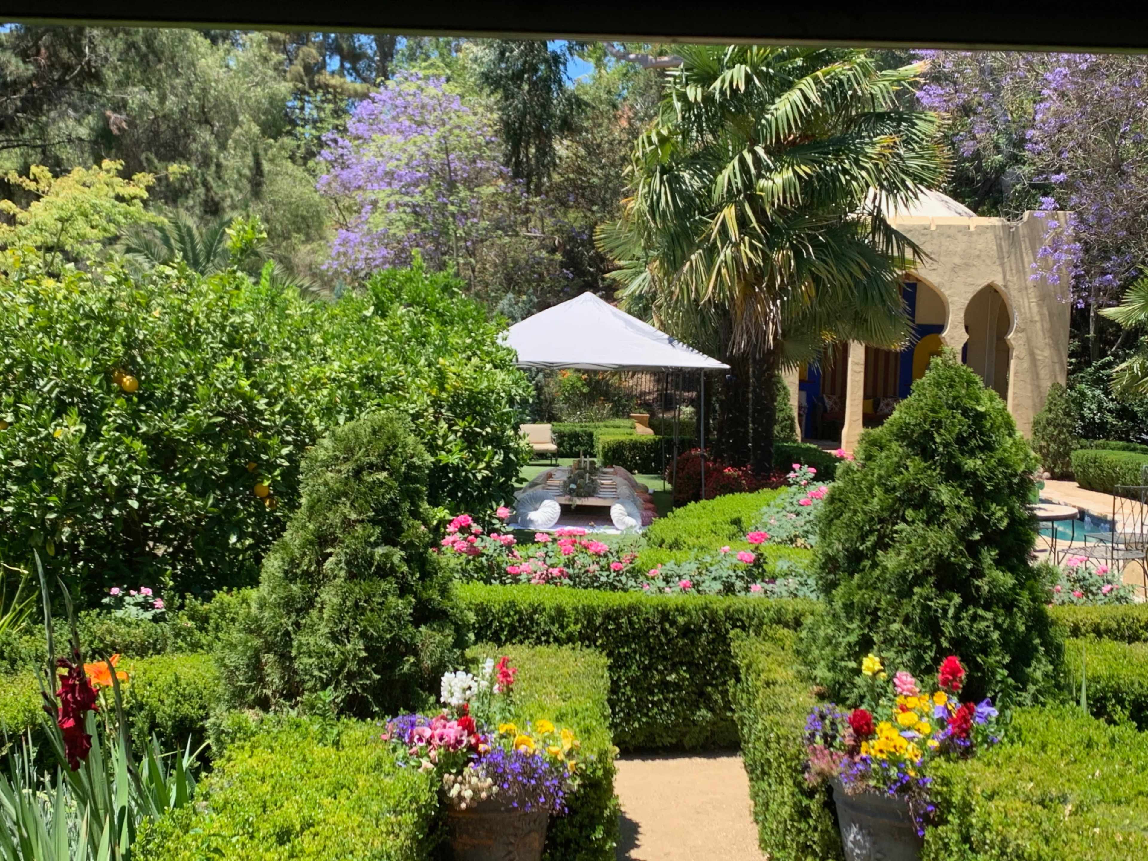 The image depicts a landscaped garden featuring colorful flowers, manicured hedges, and a shaded gazebo surrounded by lush greenery.