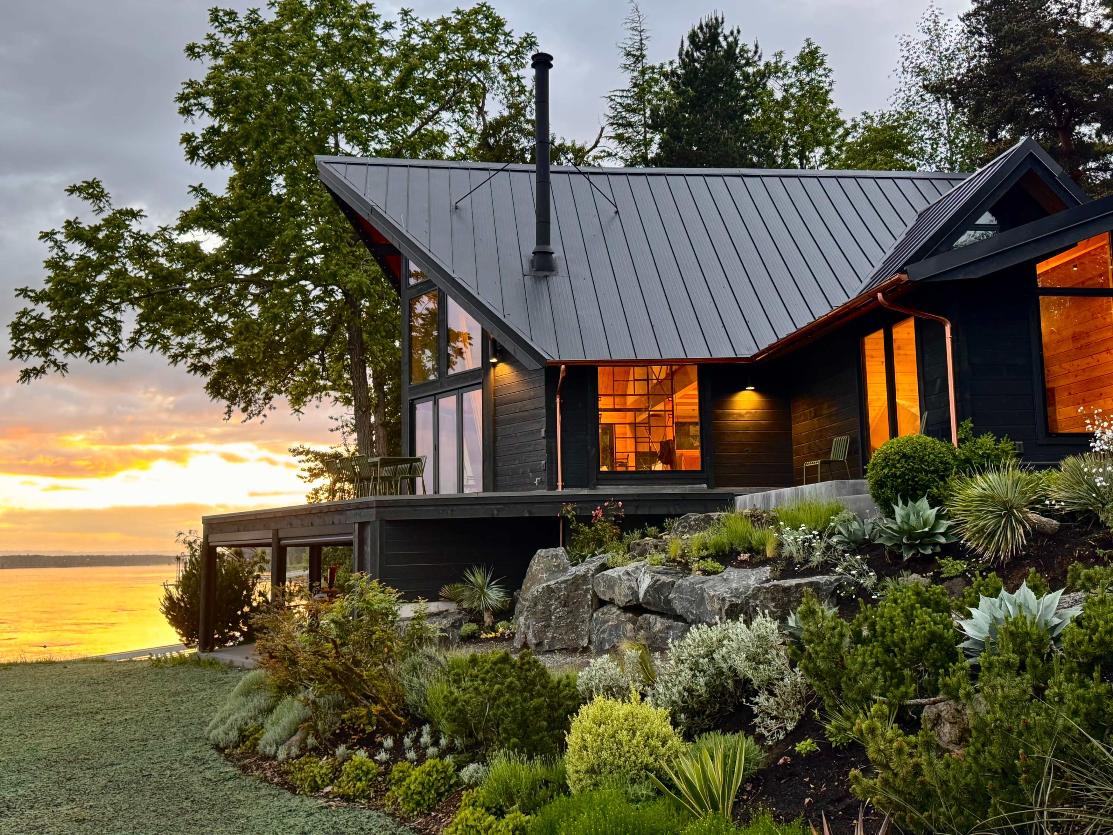 The image depicts a modern house with a slanted roof, situated on a waterfront property, surrounded by lush greenery and illuminated by a sunset in the background.