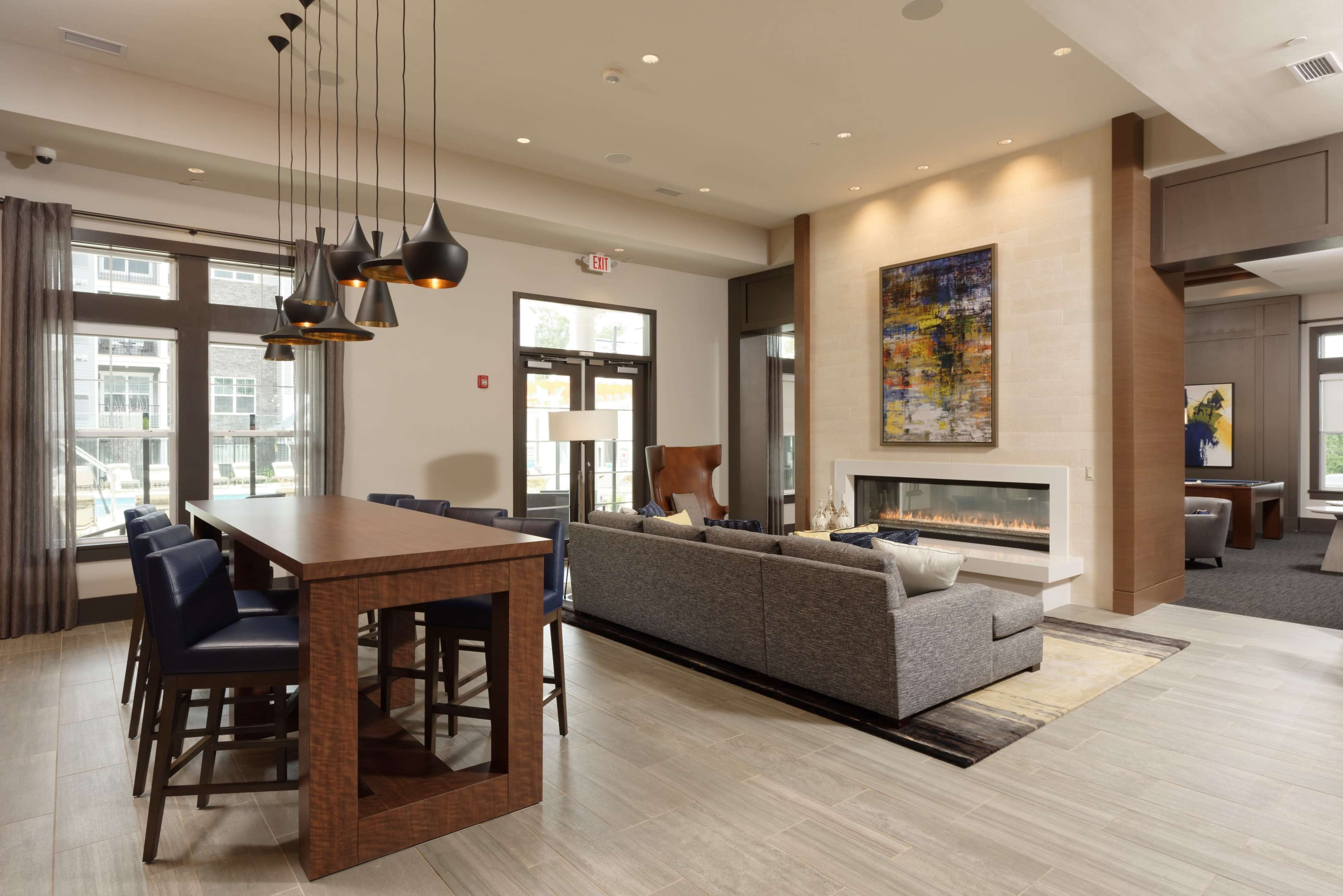A modern lobby area featuring a seating arrangement with a gray sofa, a long wooden table with high chairs, and large windows allowing natural light to fill the space.