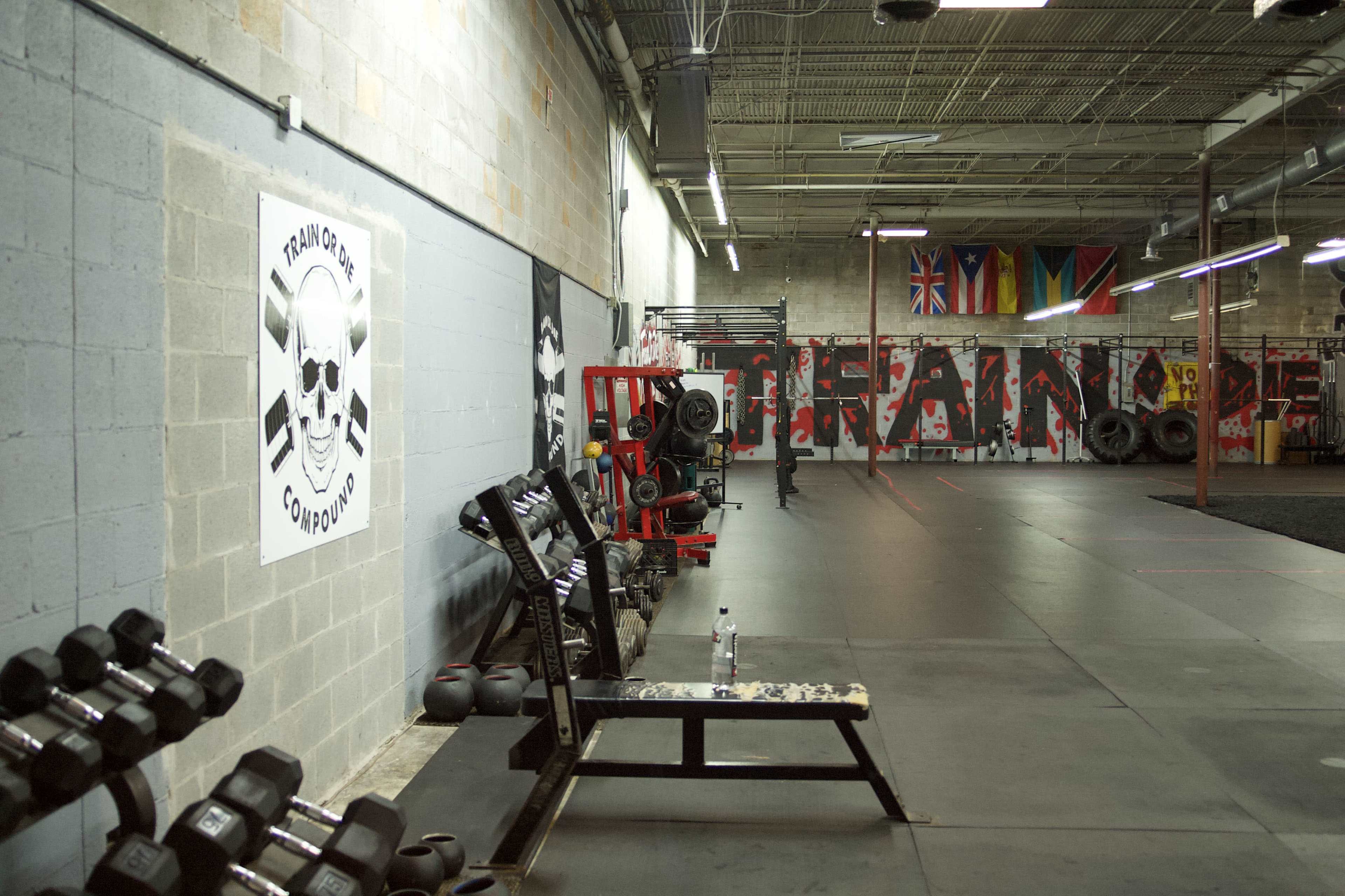 A gym interior with workout equipment, including dumbbells and benches, along with a wall featuring a large skull logo and various flags hanging from the ceiling.