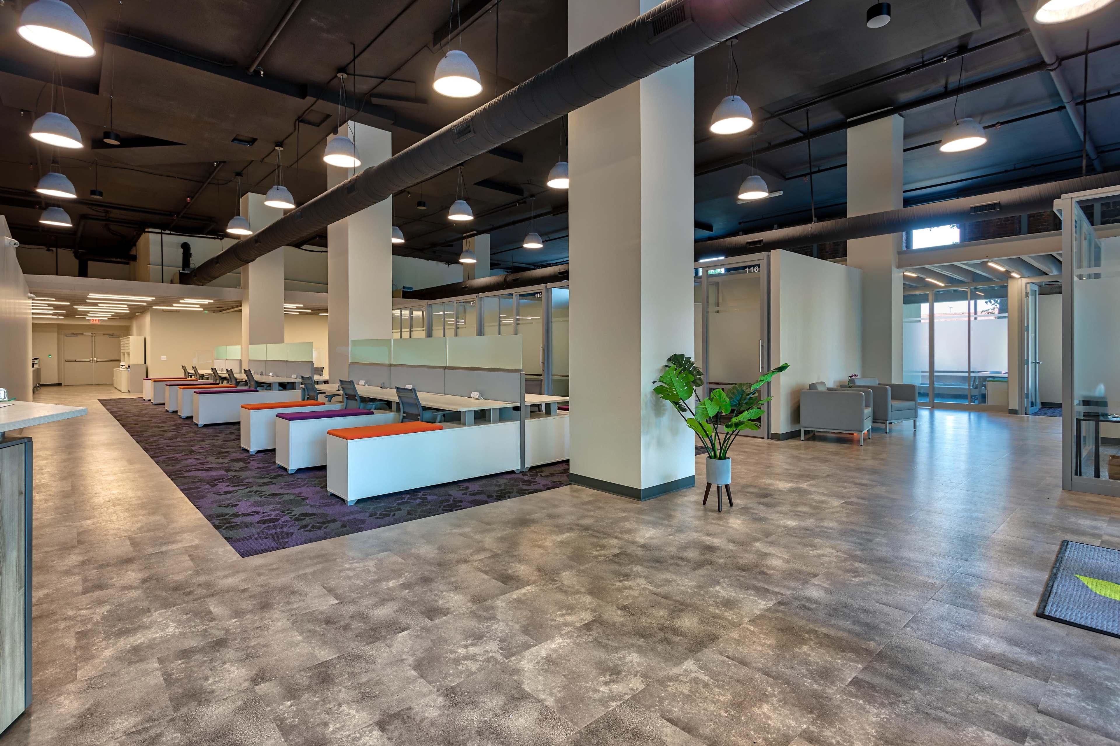 The image shows a modern office space with multiple workstations arranged in a grid layout, featuring large windows that let in natural light and an area with seating and plants.