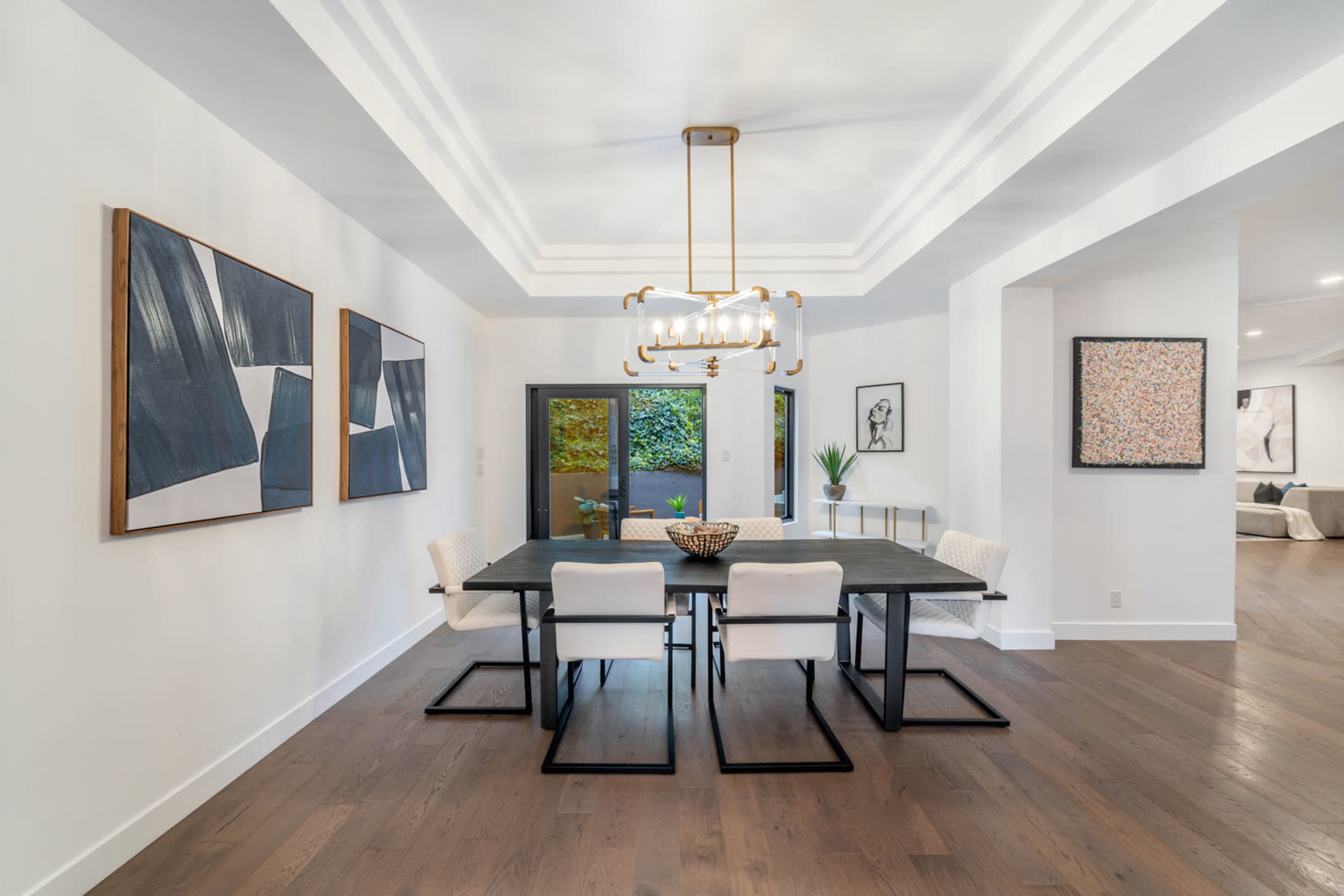 A modern dining room with a rectangular table, four white chairs, and artwork on the walls, illuminated by a stylish chandelier.