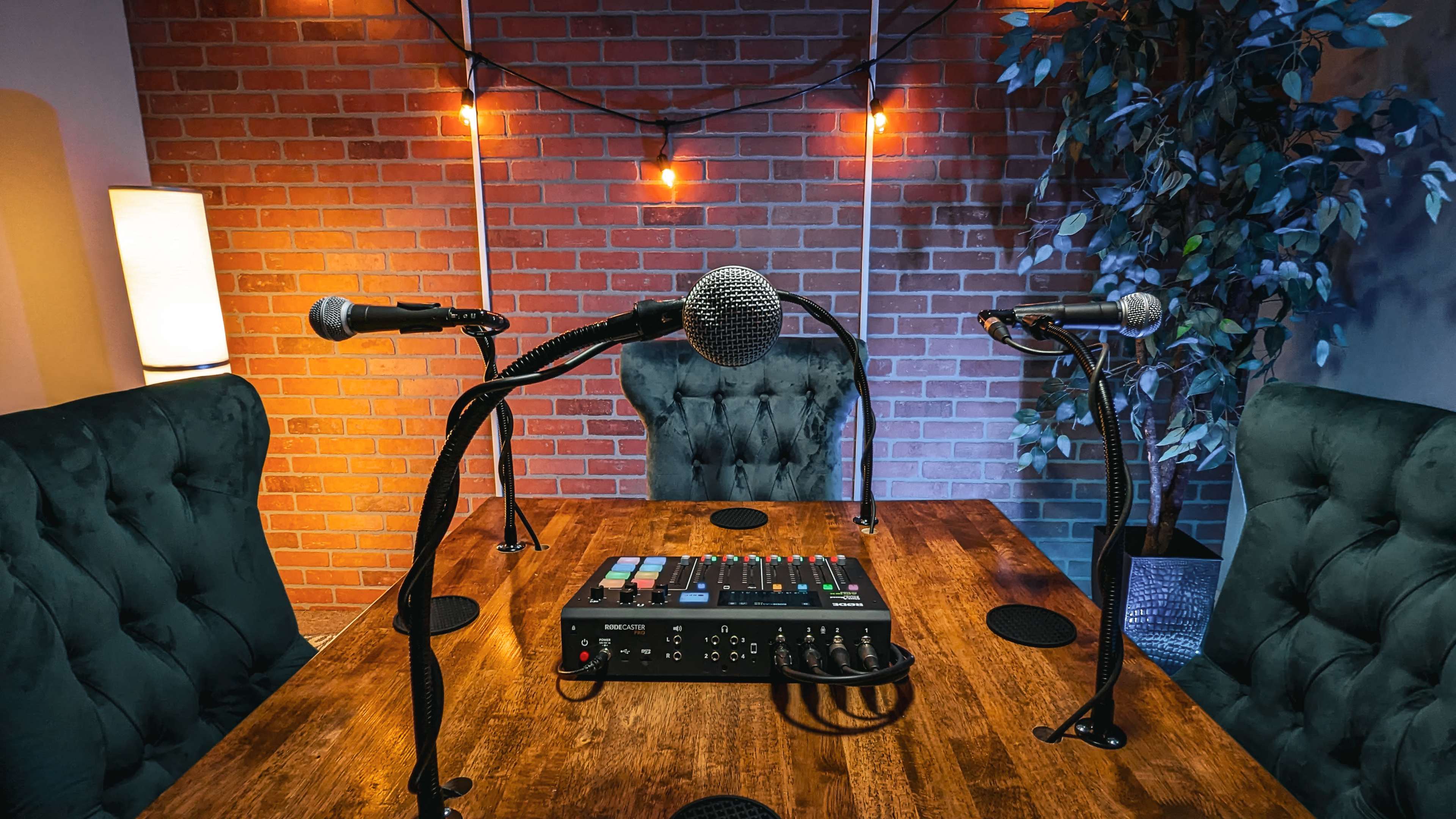 A wooden table with microphones and a sound mixer, set against a brick wall with string lights and a potted plant.