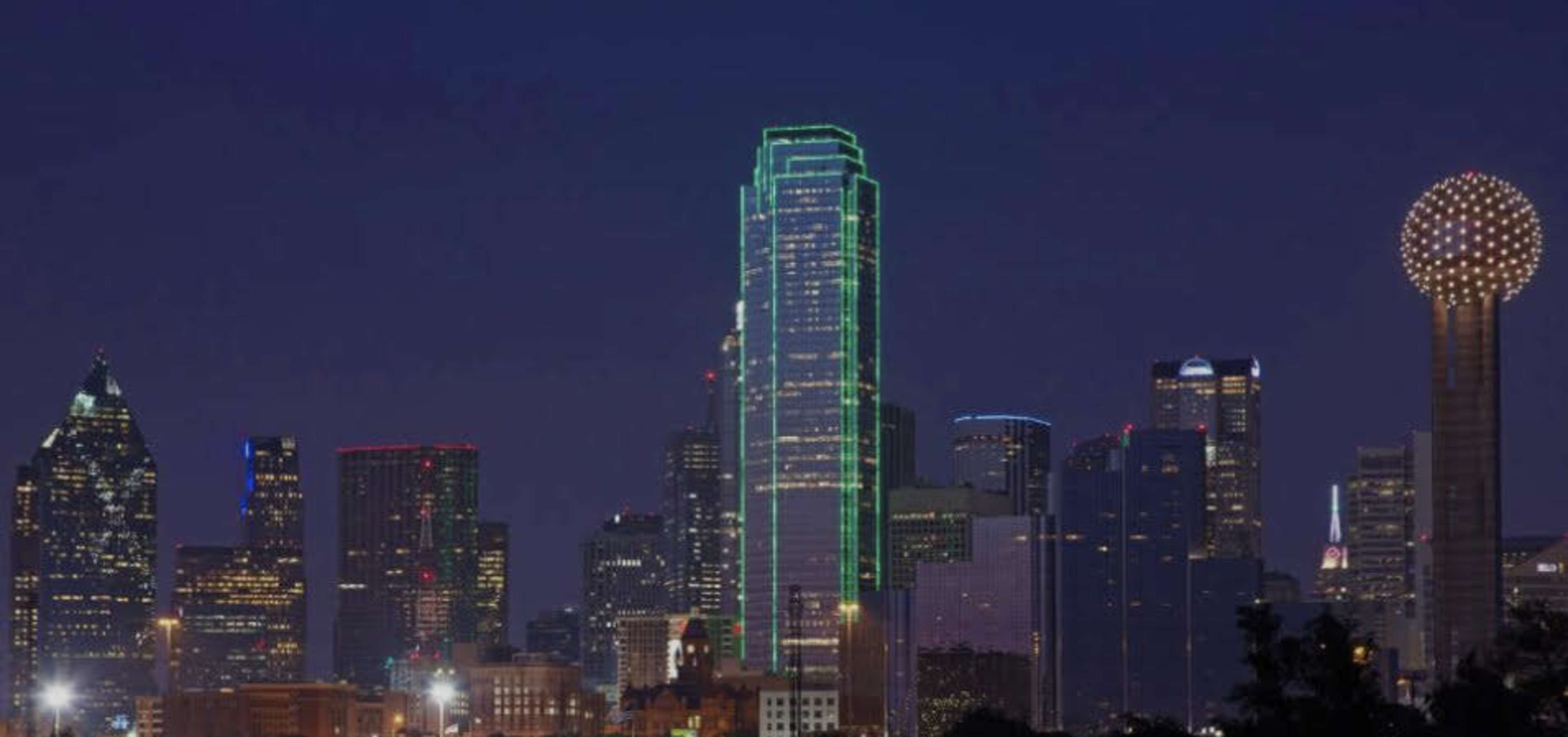 The skyline of Dallas is illuminated at night, featuring distinctive skyscrapers, including a tall building highlighted in green and the unique structure of the Reunion Tower.