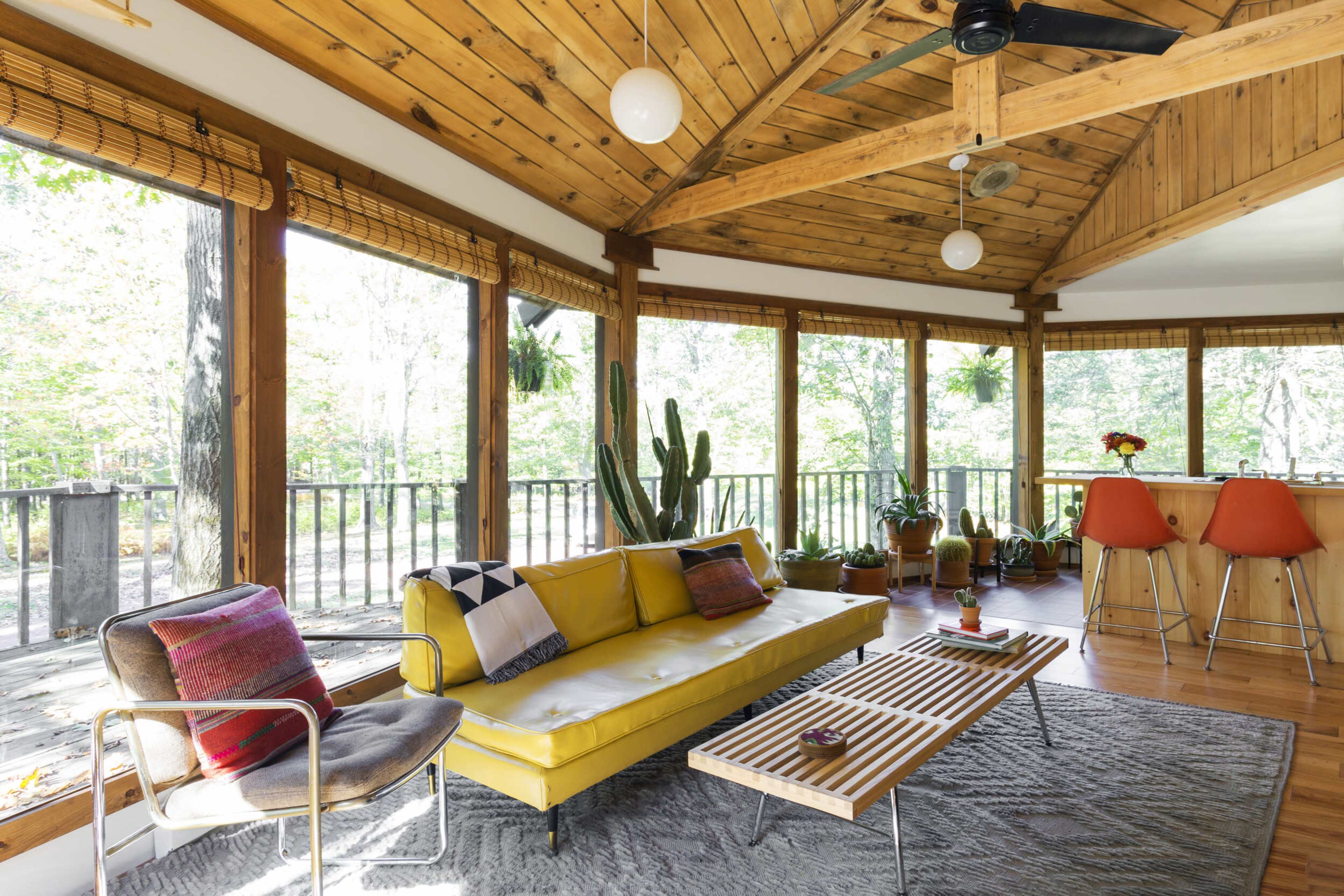 A modern living room featuring a yellow couch, a wooden ceiling with exposed beams, large windows overlooking a natural landscape, and a dining area with orange chairs.