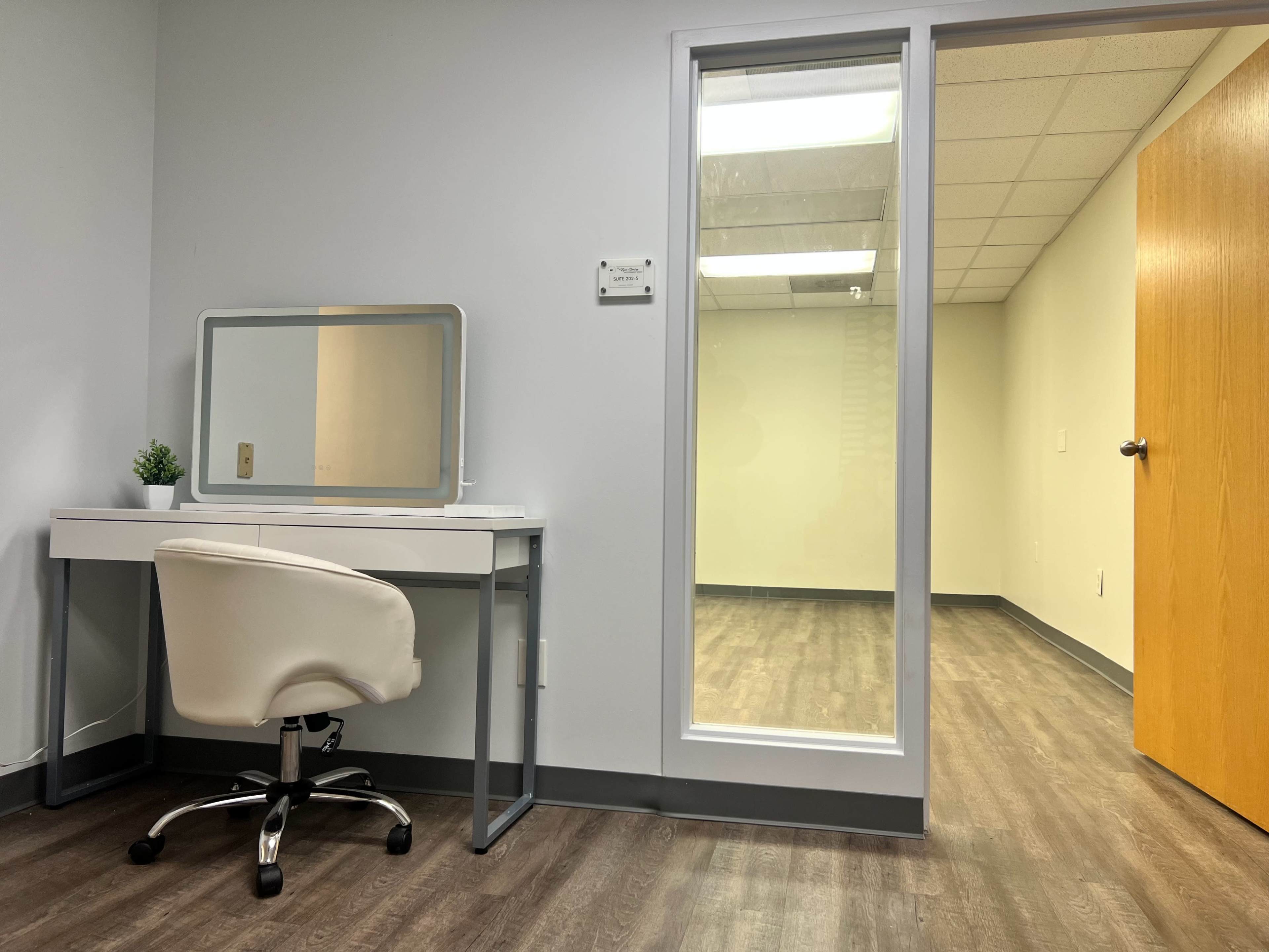The image shows a small office space with a white desk, a chair, a potted plant, and a glass-walled room in the background.